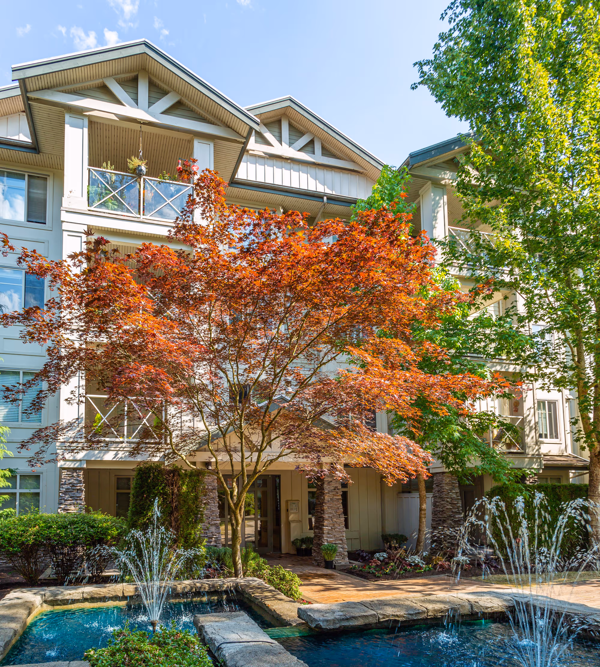 Apartment building with balconies, a red-leafed tree, and two water fountains in the foreground on a sunny day. Kumaken apartments