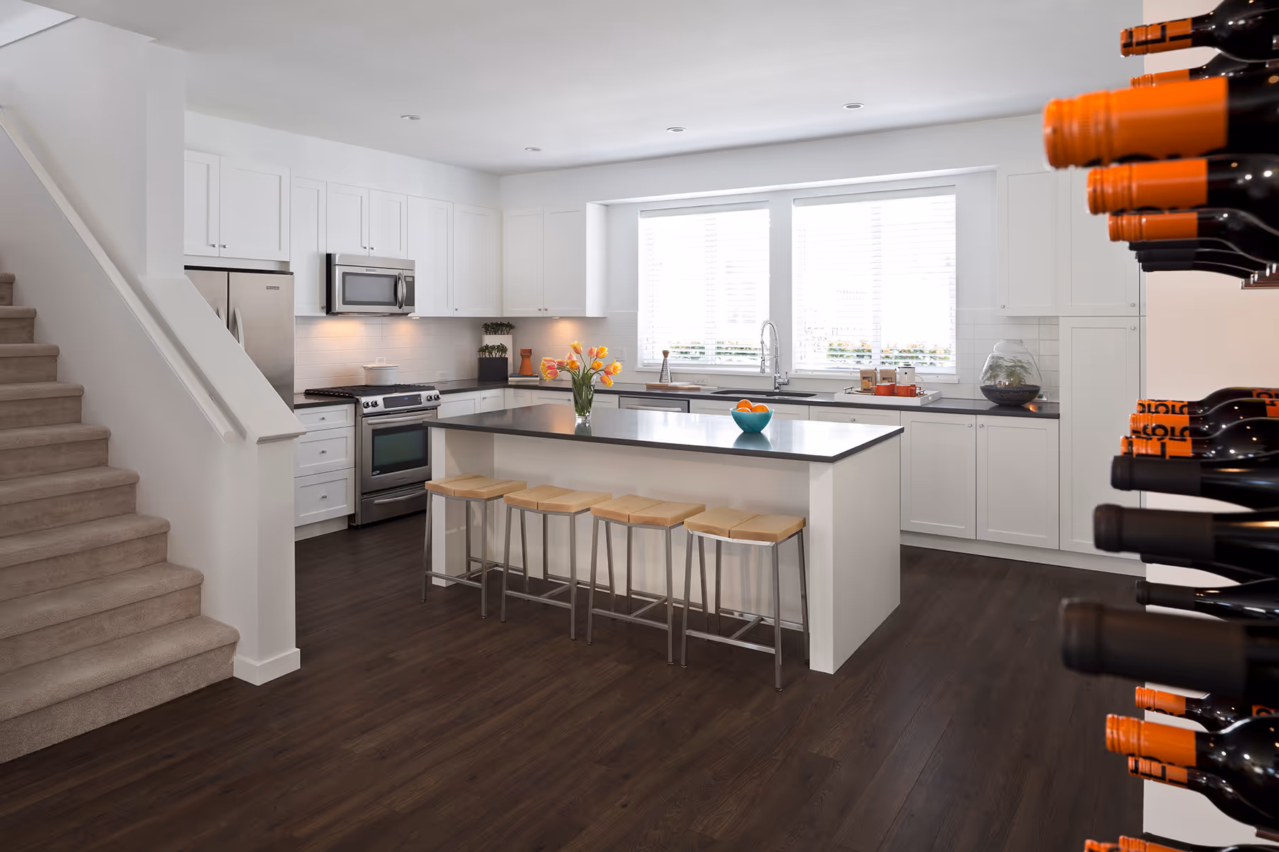 Modern kitchen with white cabinets, stainless steel appliances, a central island with four wooden stools, flowers, and a bowl of oranges, with wine bottles displayed on the right and stairs on the left by Dawson + Sawyer