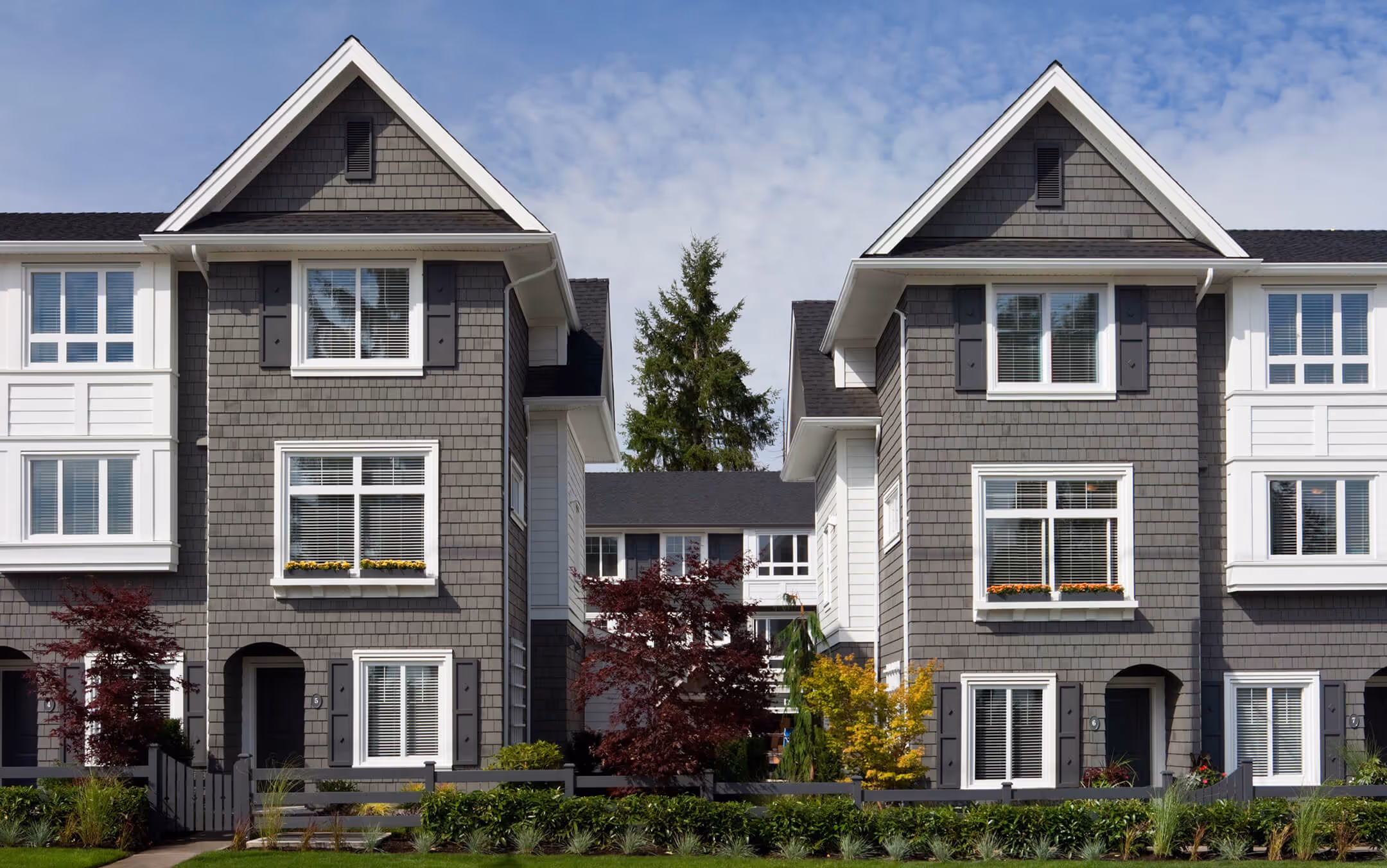 Two gray townhomes with white trim, large windows, flower boxes, and small front gardens under a partly cloudy sky. Sunnyside by Dawson + Sawyer