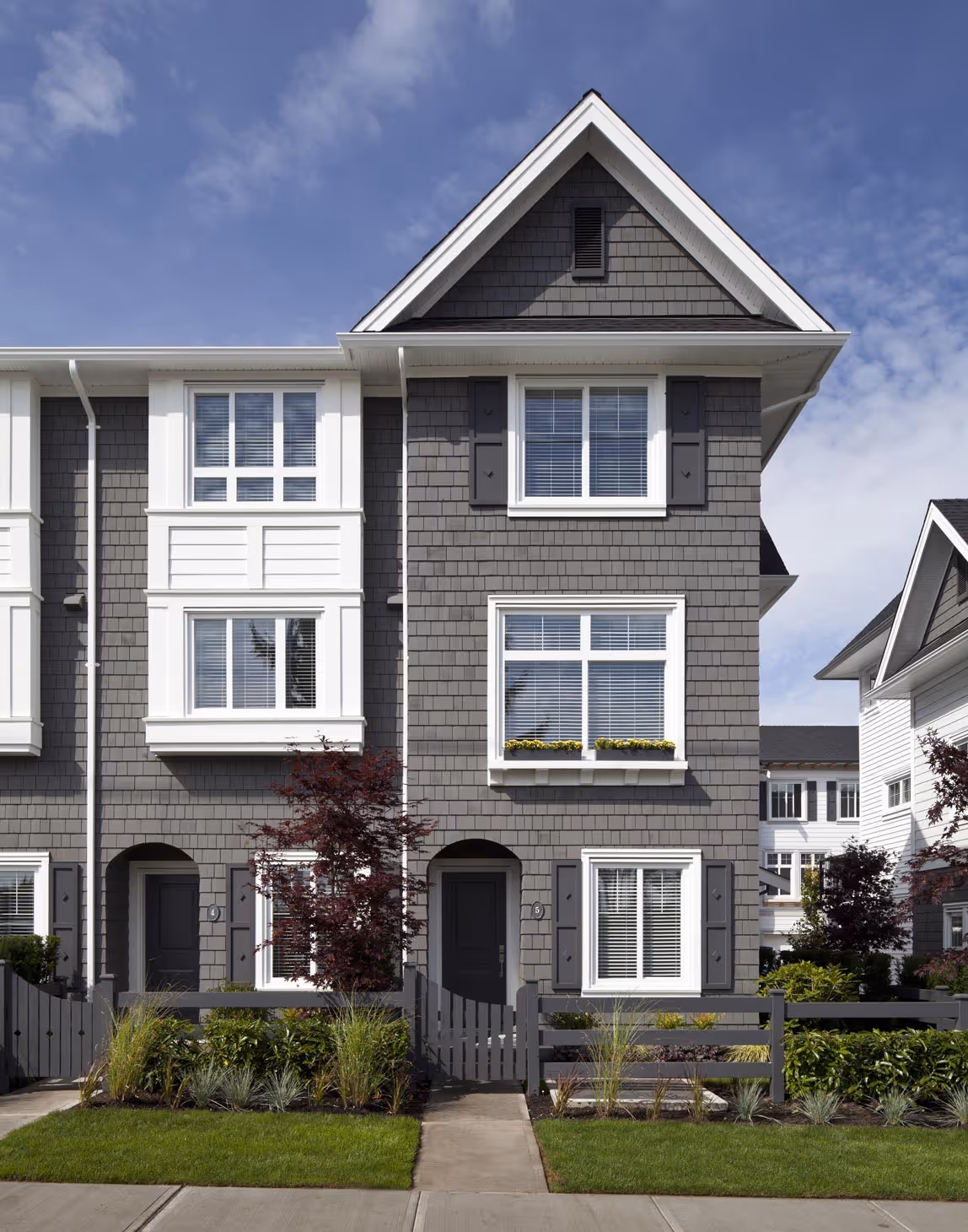 Modern grey townhome with white trim, front doors, windows with shutters, and well-maintained front gardens under a blue sky.