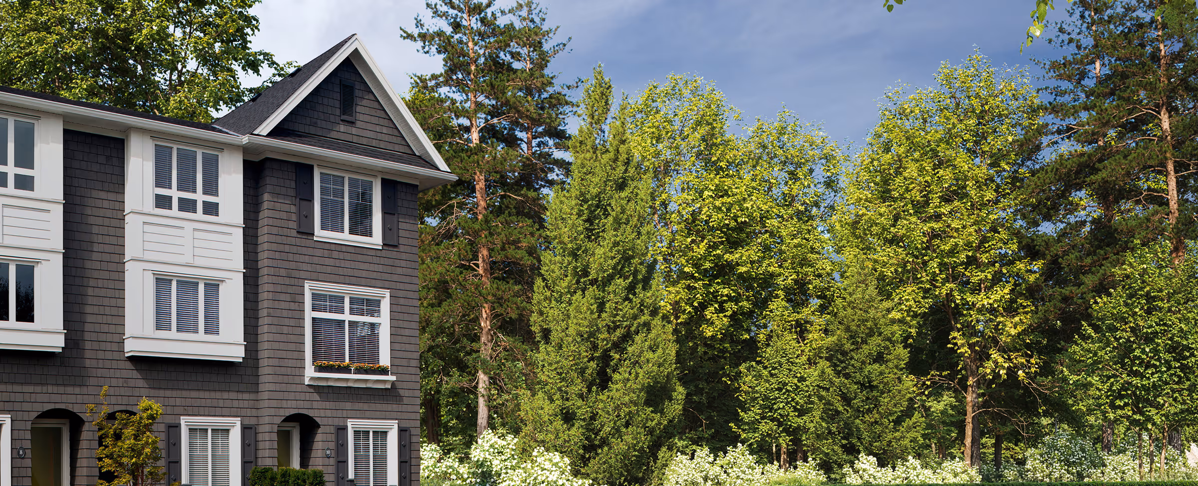 Row of gray townhomes at Queens in bear creek with white trim next to a lush green garden and tall trees under a partly cloudy sky.