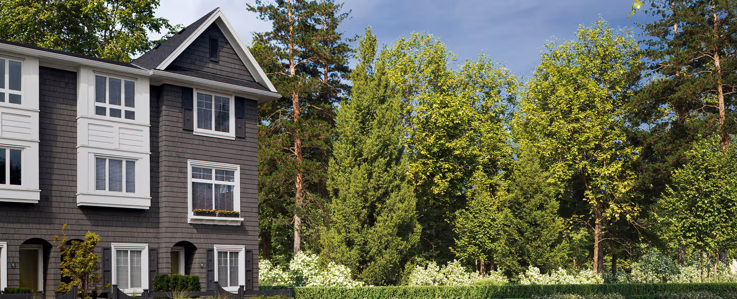 Row of gray townhomes at Queens in bear creek with white trim next to a lush green garden and tall trees under a partly cloudy sky.