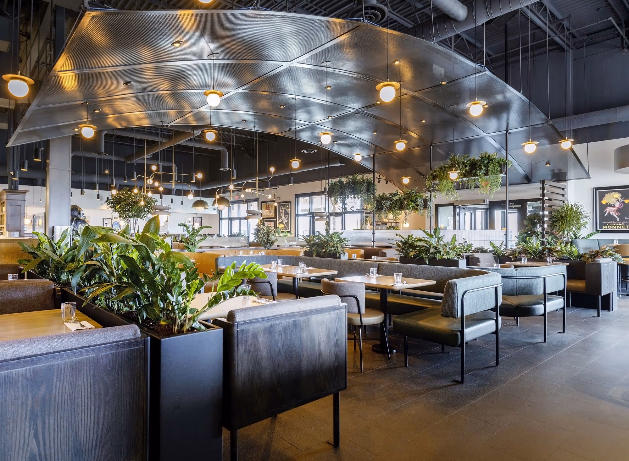 Modern restaurant interior with cushioned booth seating, wooden tables, green plants, and hanging pendant lights under a metallic ceiling panel.