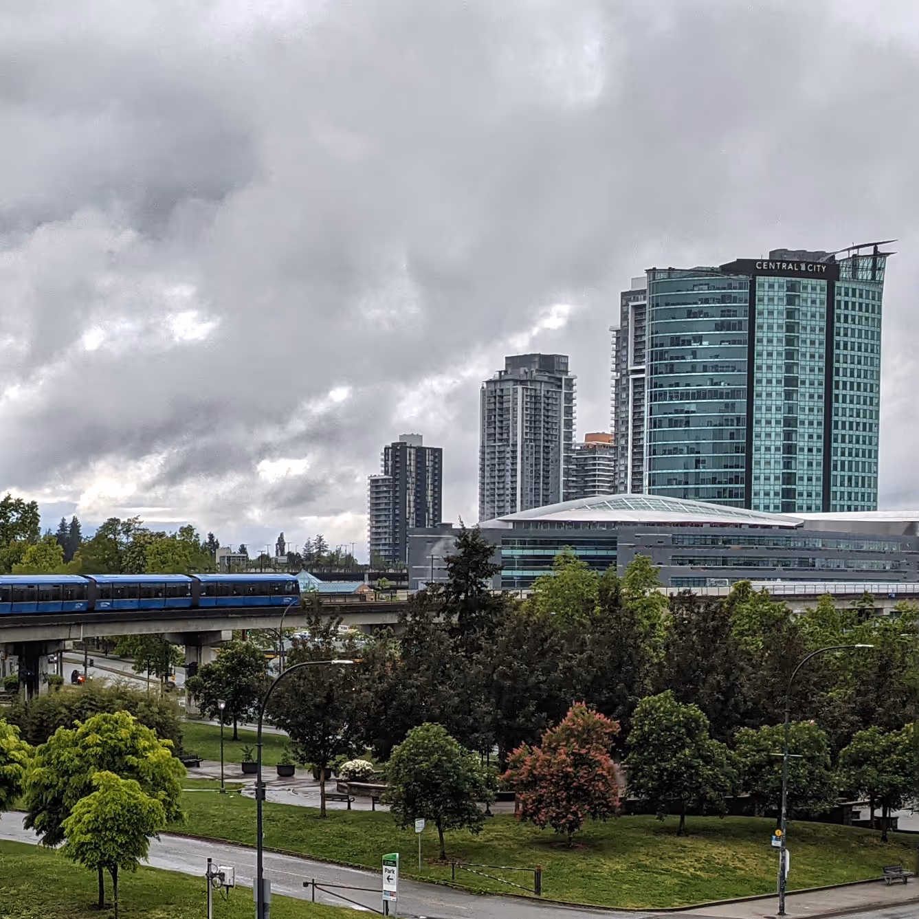 Urban cityscape with modern high-rise buildings, a blue elevated train on tracks, and green park trees under a cloudy sky.