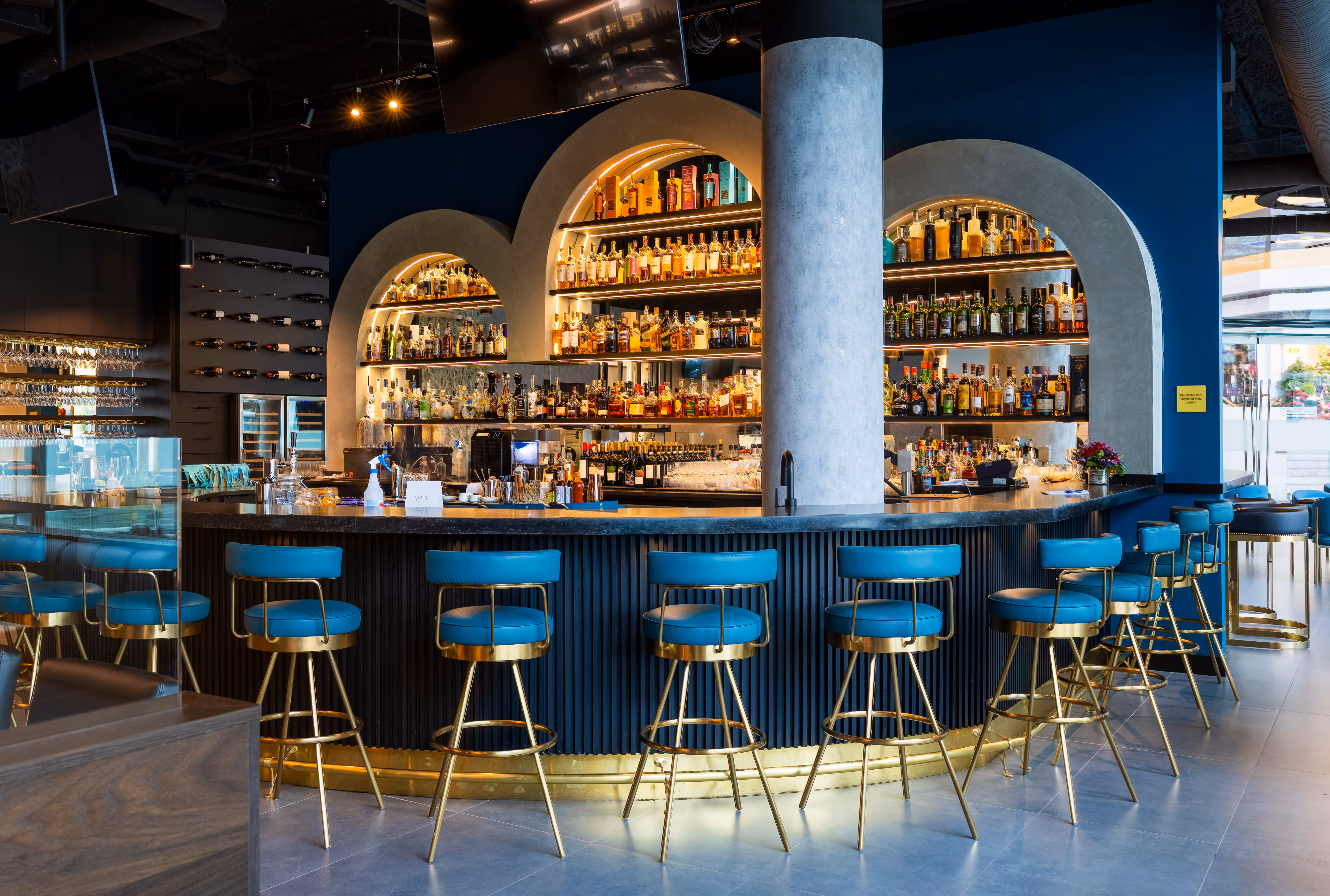 Modern bar with blue and gold stools lined up around a curved counter and illuminated shelves stocked with bottles.