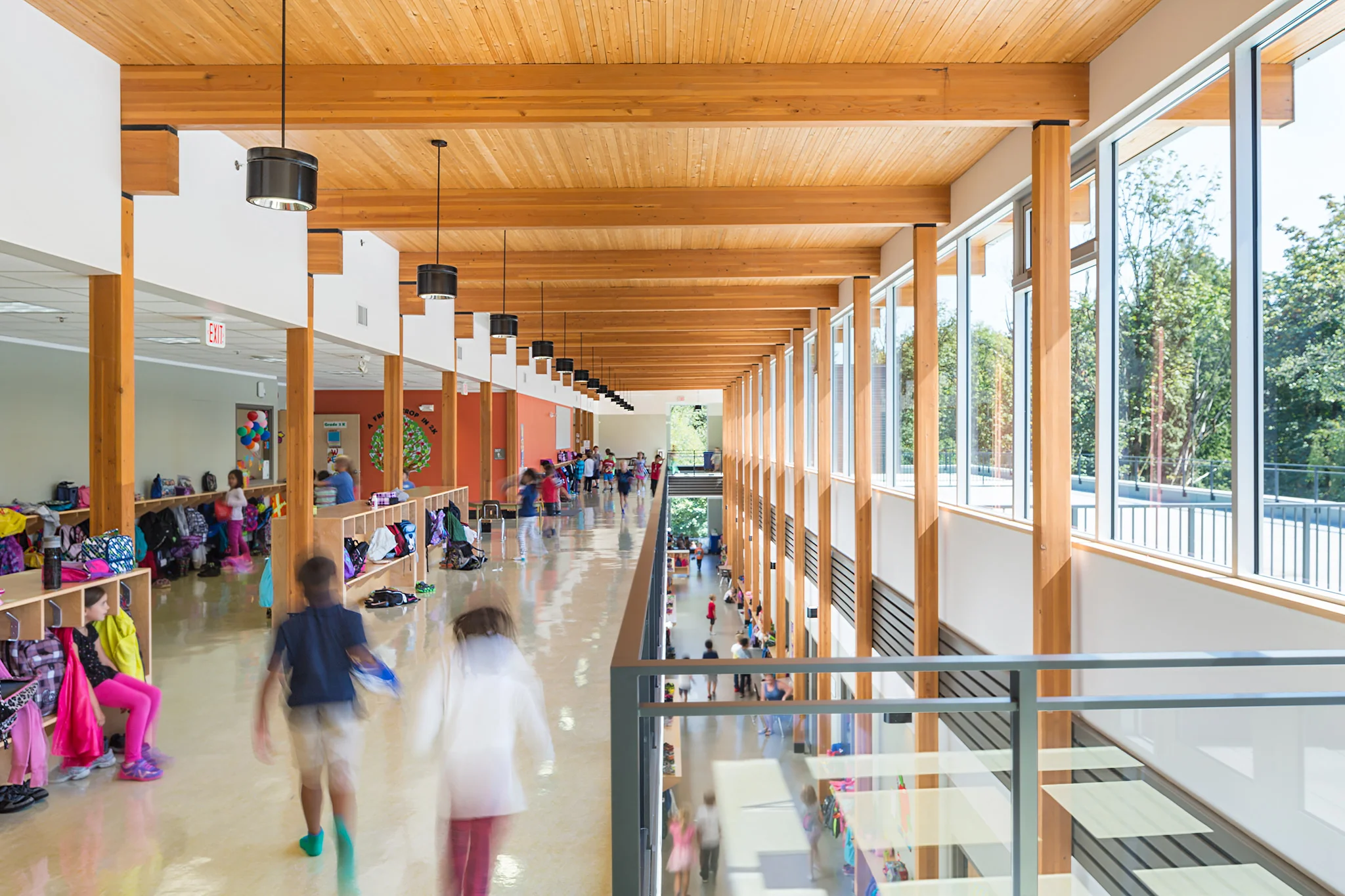 Bright, spacious school hallway with large windows, wooden beams, and children walking and sitting.