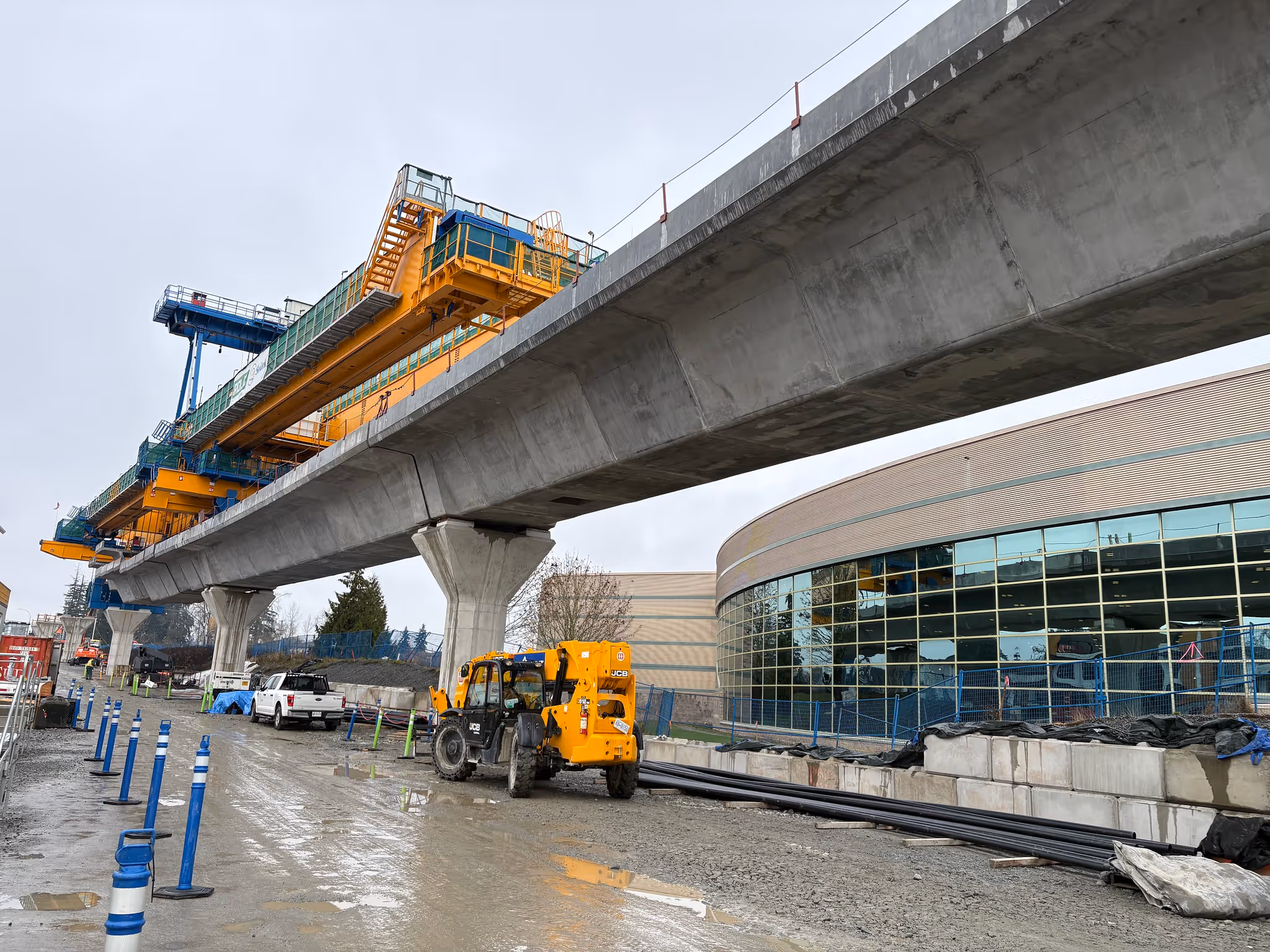 Elevated concrete bridge under construction with heavy machinery and vehicles on muddy ground next to a modern building with large windows.