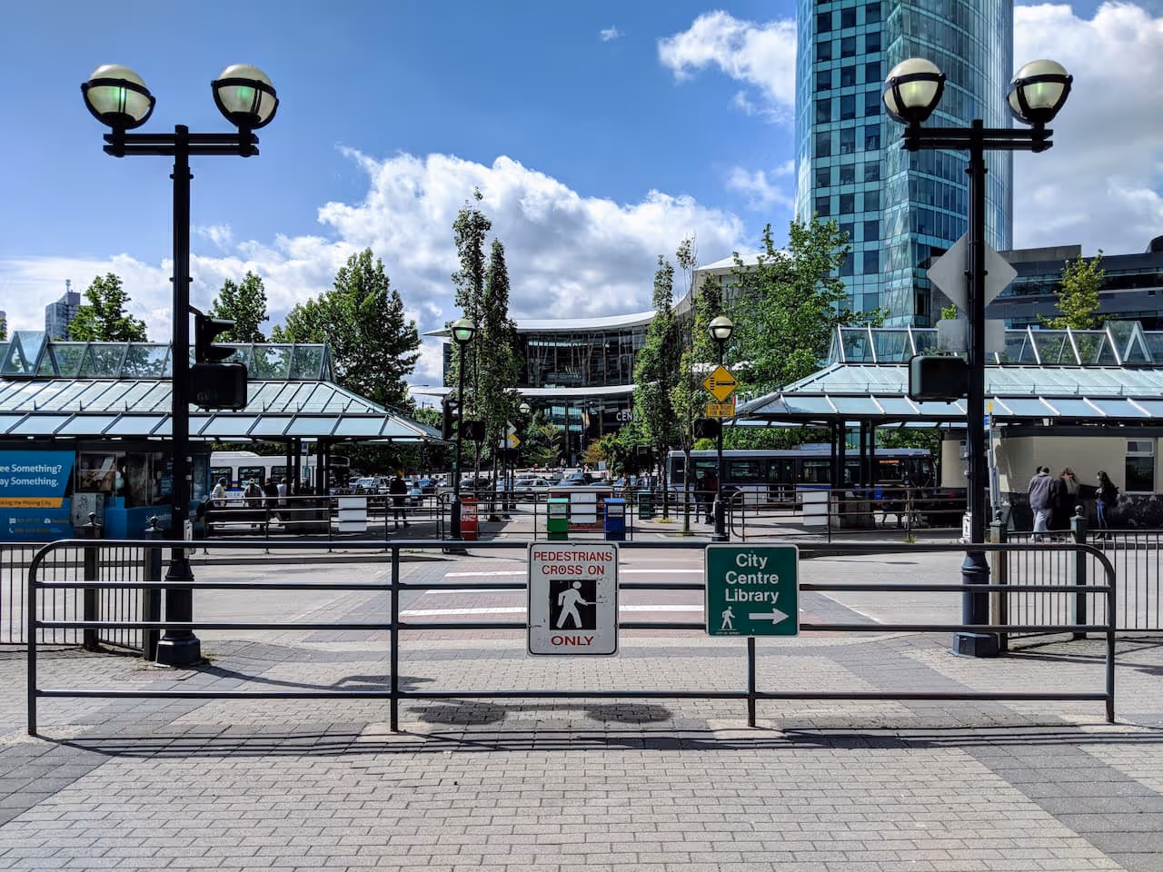 Urban bus exchange with glass-covered bus shelters, pedestrian crossing signs, tall trees, and modern buildings under a partly cloudy sky.