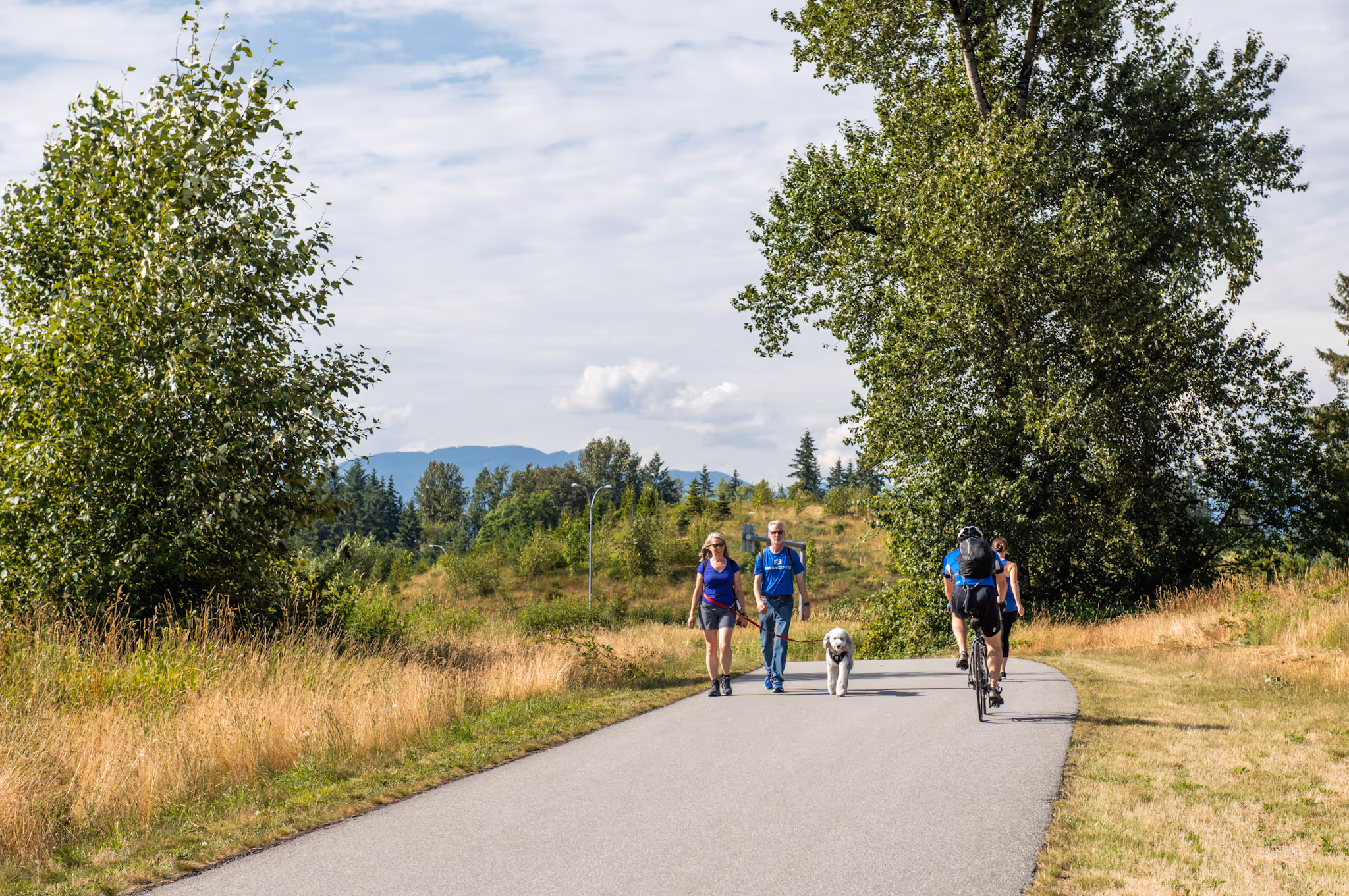 A couple walking a dog and two cyclists on a paved trail surrounded by trees and grass under a partly cloudy sky.