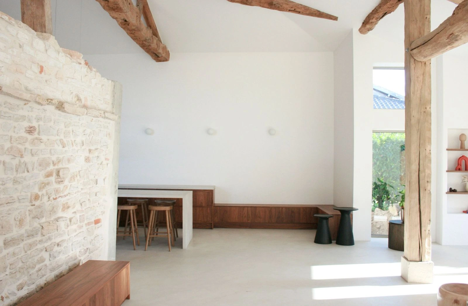 Pièce minimaliste avec poutres en bois apparentes, murs blancs, mur en pierre, tabourets en bois, banc intégré en bois et lumière naturelle.