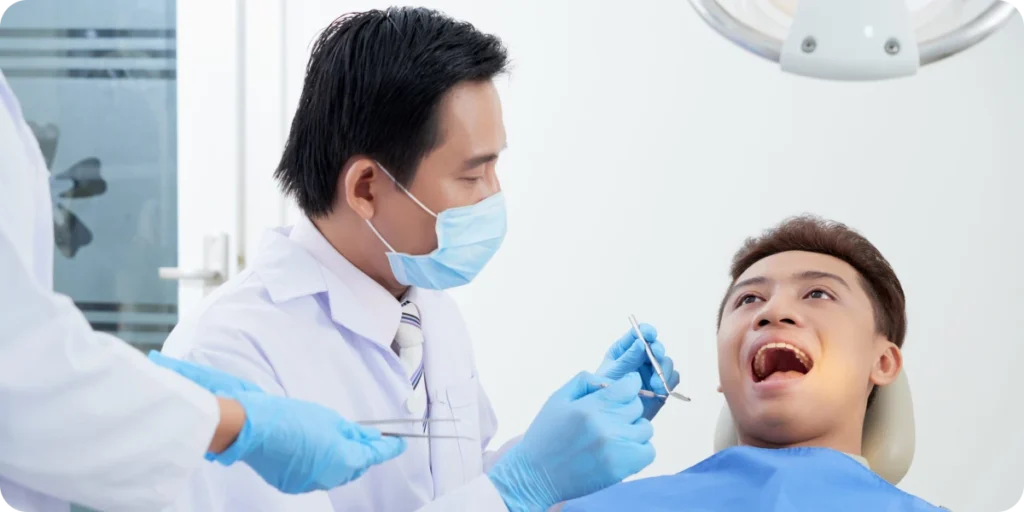 A clinical treatment room where a healthcare professional in a white coat and blue medical gloves prepares dental or medical instruments while interacting with a patient seated in a treatment chair. Another medical staff member stands nearby holding additional tools. The bright, sterile environment includes white walls, a large overhead examination light, and modern clinic equipment. The scene represents medical device usage, clinical procedures, regulatory compliance, and contexts relevant to NMPA requirements such as post‑market surveillance, clinical safety, and authorized agent responsibilities.