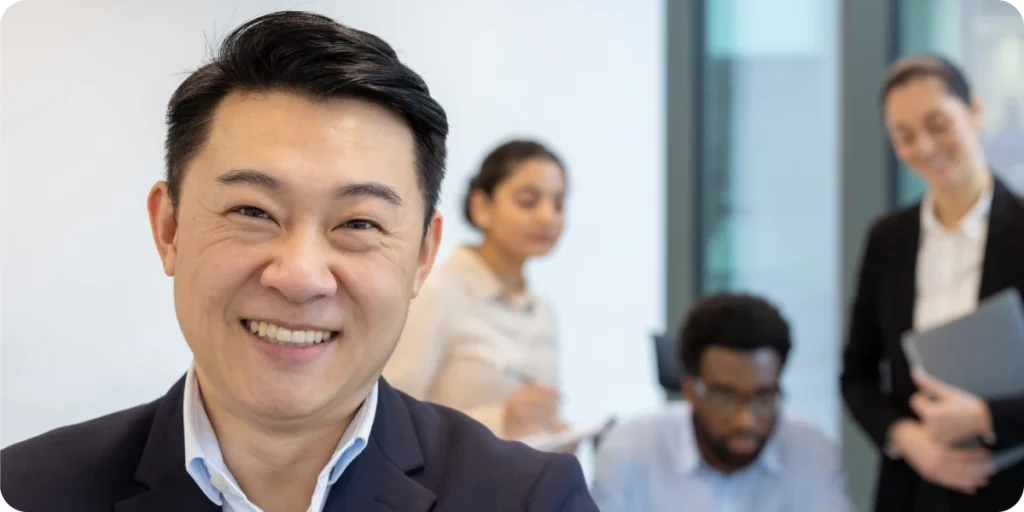 A professional office environment featuring several business professionals standing and sitting in a meeting area. The person in the foreground is positioned close to the camera, dressed in formal business attire. In the background, additional team members are gathered near a table, with documents and a laptop visible, suggesting regulatory consulting or compliance discussions. The modern office has bright lighting and large glass panels. The scene conveys a corporate setting related to regulatory affairs, medical device compliance, and responsibilities typical of an NMPA legal agent, such as product registration oversight, documentation management, and regulatory liaison activities.