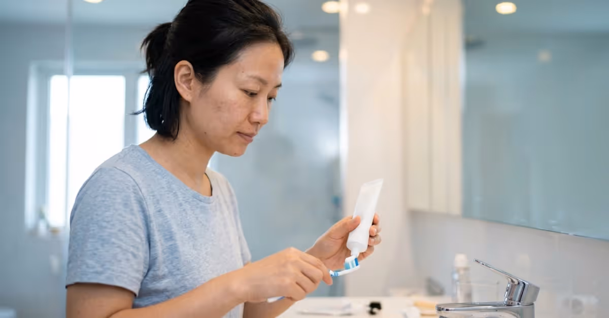 Person standing at a bathroom sink examining a cosmetic tube and applying a small amount of cream to a fingertip, suggesting a skincare routine or product check in a clean, modern setting; relevant to consumer product use and safety evaluation.