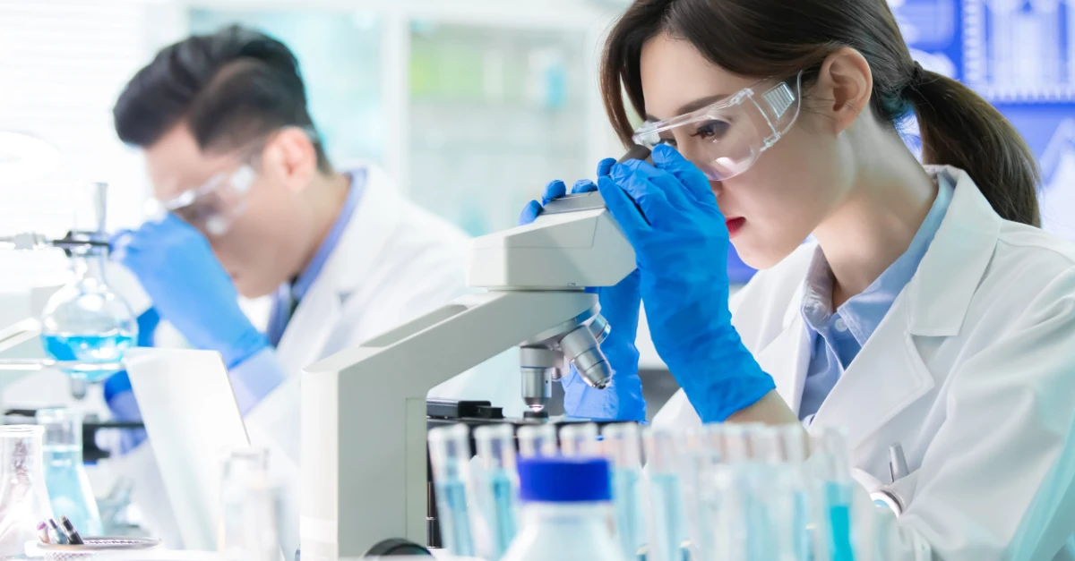 A brightly lit laboratory scene shows two scientists conducting research. In the foreground, a woman wearing clear safety goggles, blue nitrile gloves, and a white lab coat peers into a microscope with focused concentration.