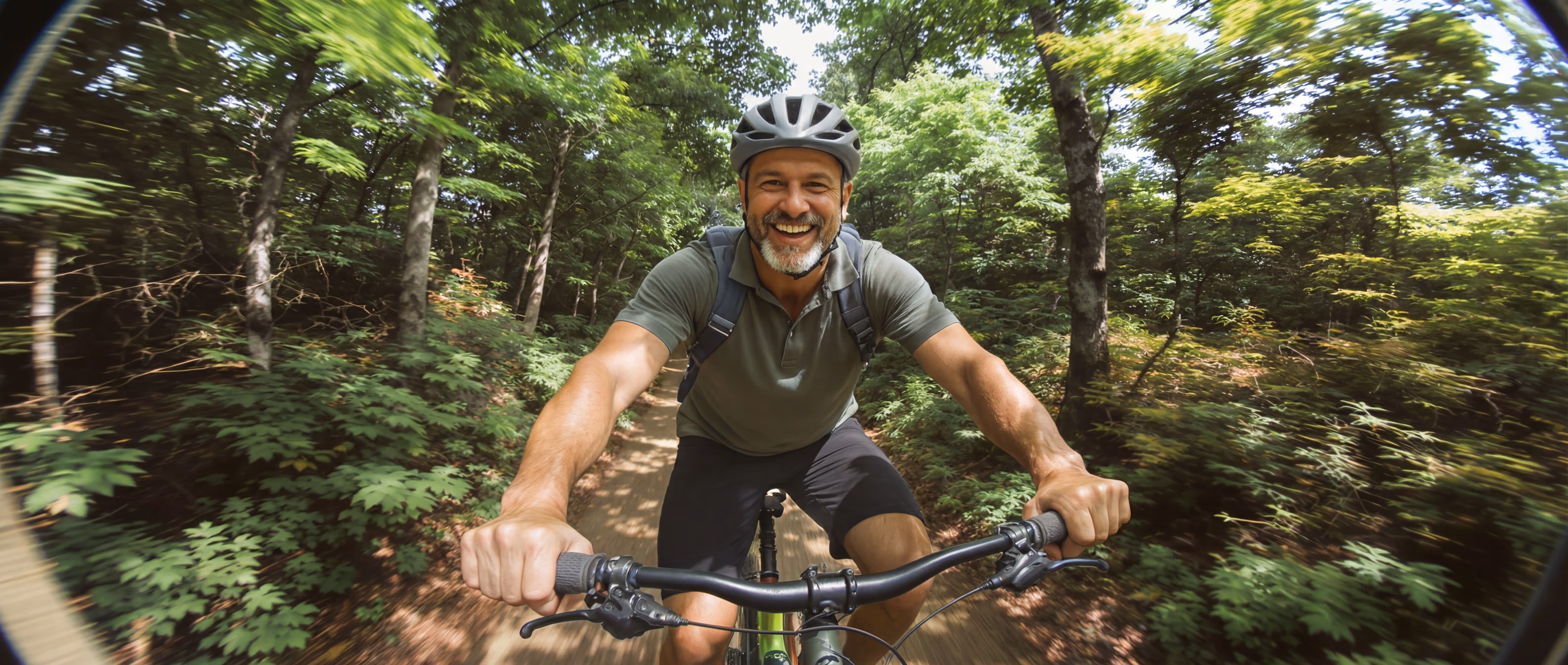 Smiling man wearing a helmet riding a bicycle on a forest trail surrounded by green trees.