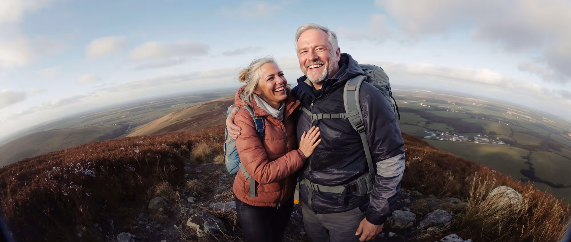 Happy middle-aged couple wearing jackets and backpacks hugging on a hilltop overlooking a wide rural landscape under a partly cloudy sky.