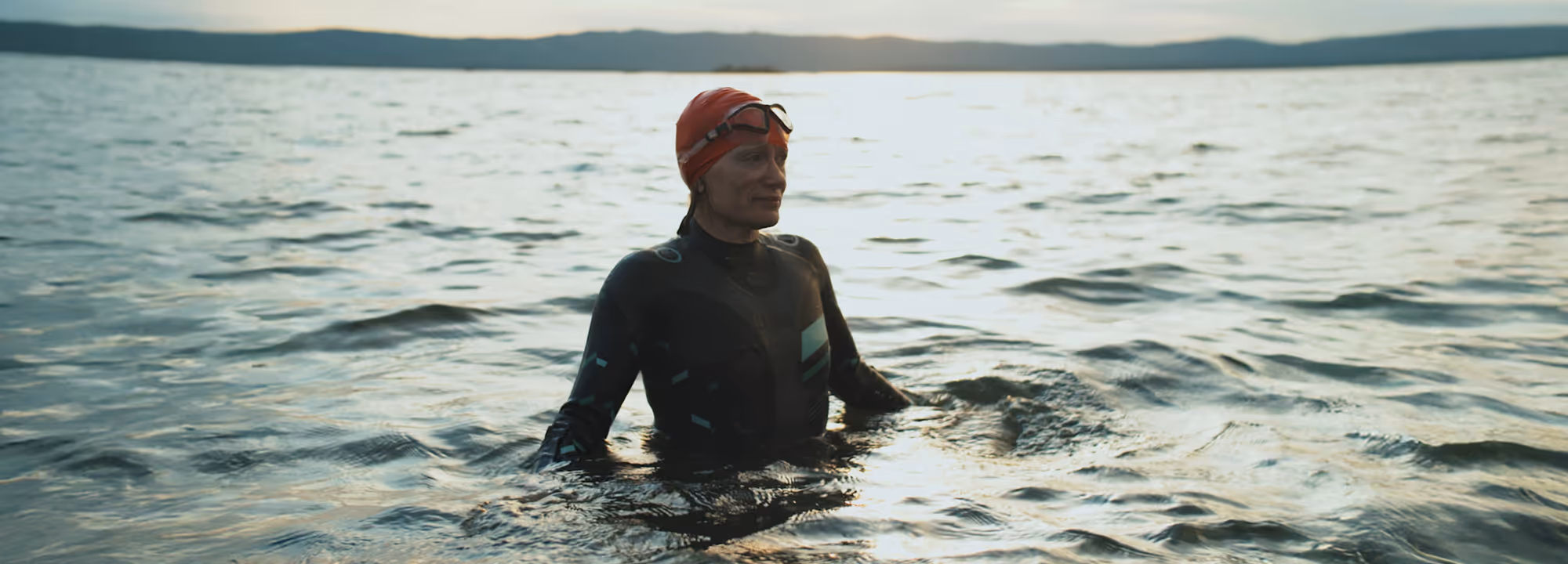 Person wearing a red swim cap and black wetsuit standing waist-deep in a calm body of water with distant hills on the horizon.