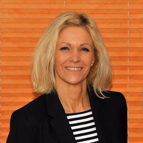 Porträt einer lächelnden Frau mit schulterlangem blondem Haar, gestreiftem Oberteil und schwarzem Blazer vor orangefarbenem Hintergrund.