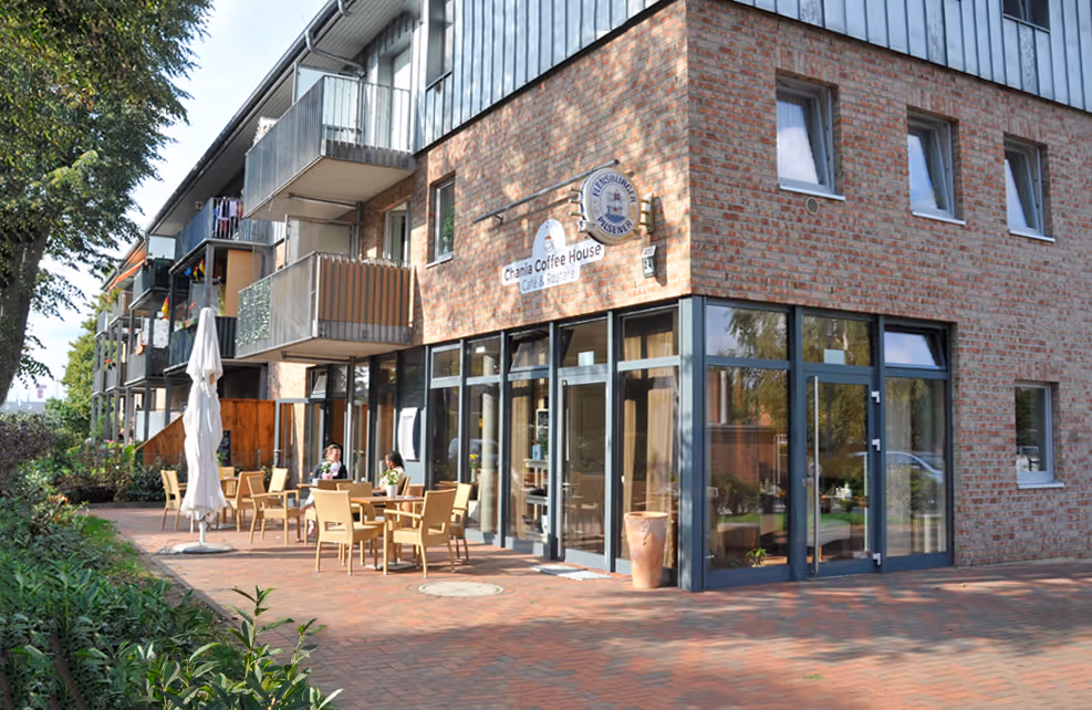 Außenbereich eines Cafés mit Terrassenmöbeln vor einem modernen Wohngebäude aus rotem Backstein.