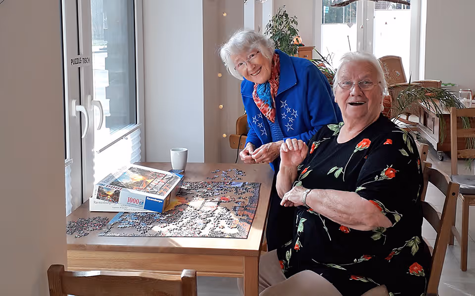 Zwei ältere Frauen sitzen an einem Tisch und arbeiten gemeinsam an einem 1000-teiligem Puzzle bei Tageslicht.