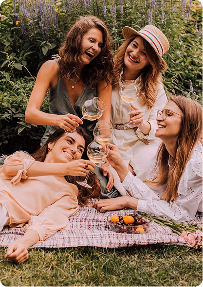 Quatre amies souriantes trinquant avec des verres de vin blanc lors d'un pique-nique sur une couverture à l'extérieur.