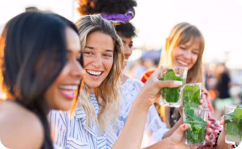 Groupe de quatre femmes souriantes portant des boissons vertes dans des verres, en plein air.