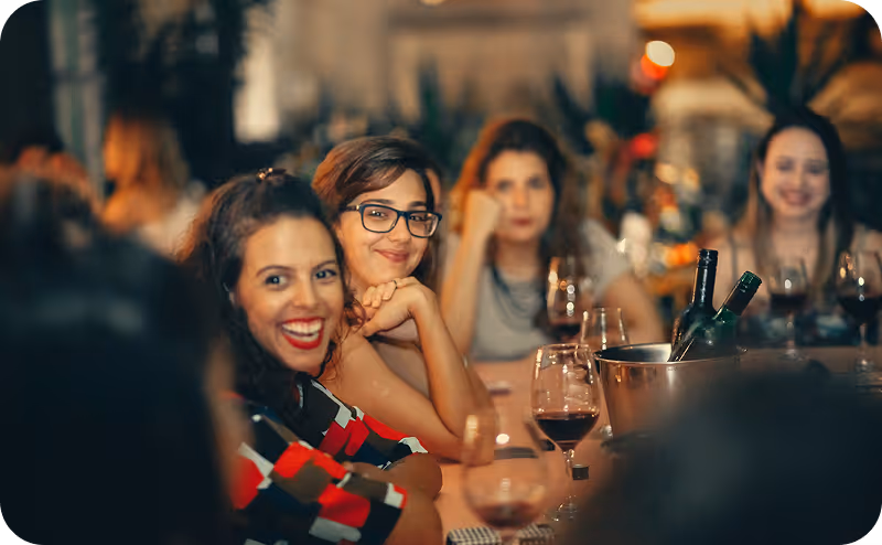 Quatre femmes assises à une table de restaurant avec des verres de vin rouge et un seau à glace contenant des bouteilles.