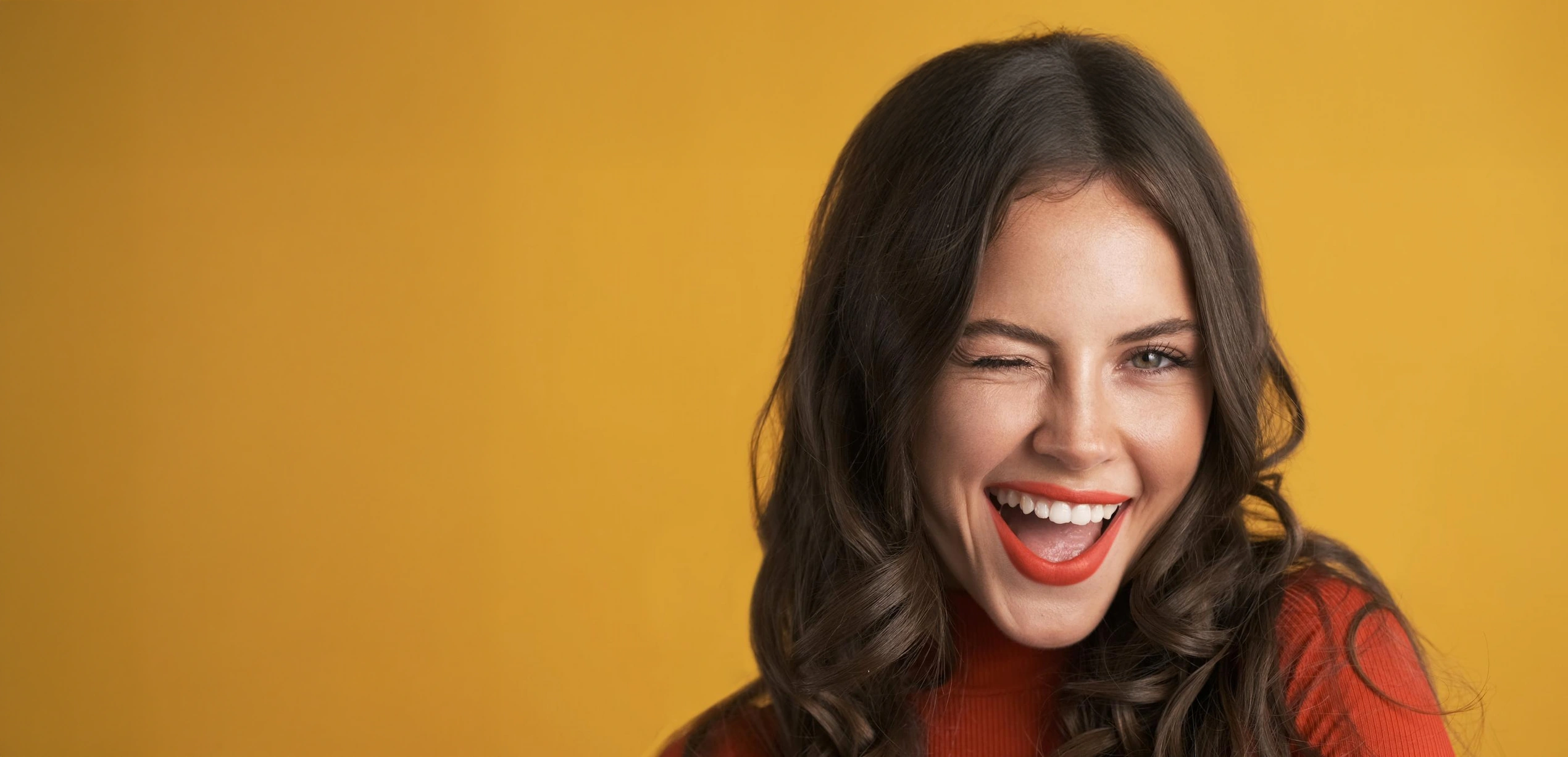 Jeune femme aux cheveux bruns bouclés, portant un pull rouge, souriant et clignant de l'œil sur un fond jaune.