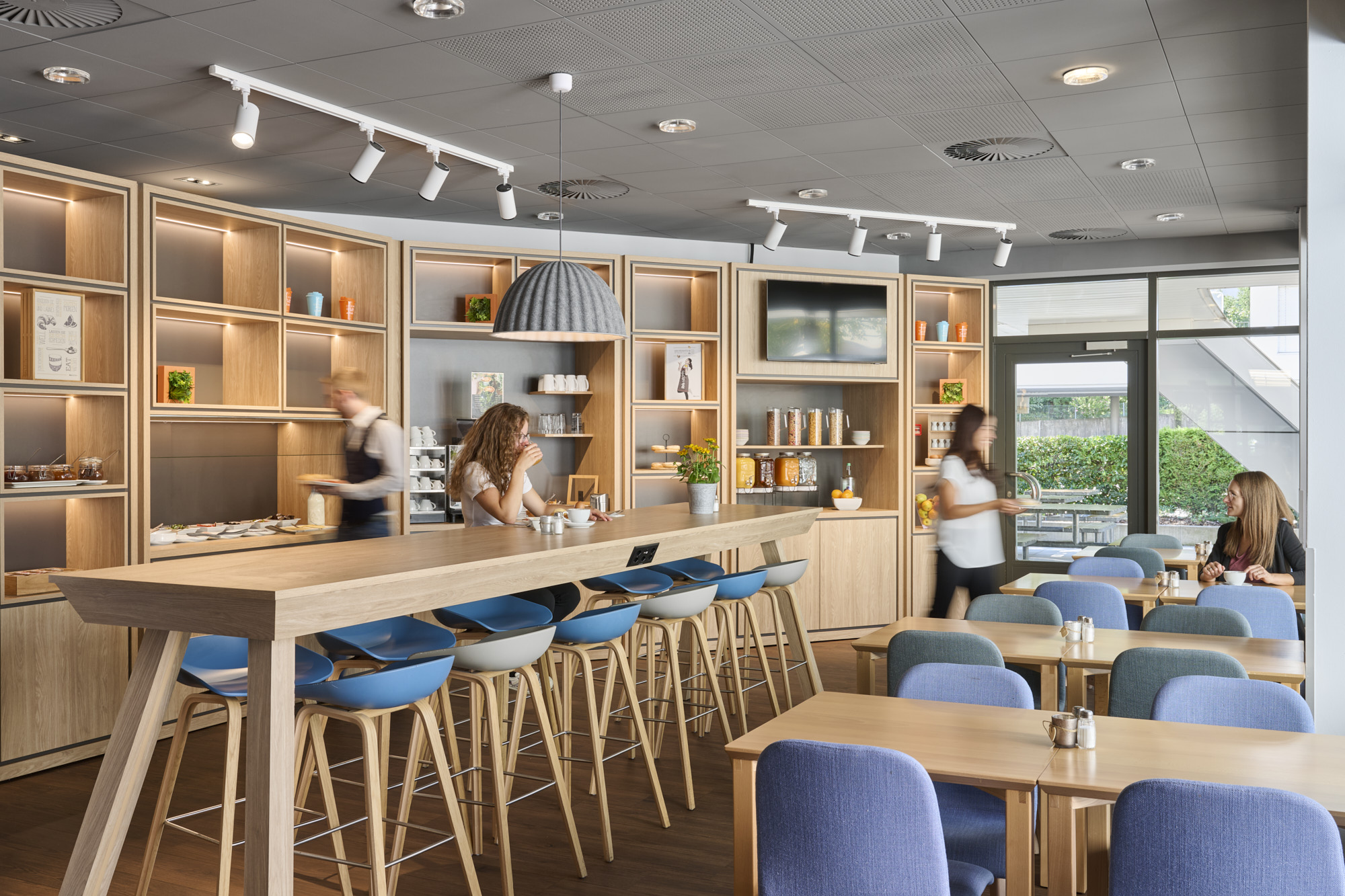 Modernes Café im Ibis Hotel in Chur mit langem Holztisch und blauen sowie grauen Hockern, weiteren Holztischen mit blauen Stühlen, Frau am Tresen beim Kaffeetrinken und Personal beim Bedienen der Gäste