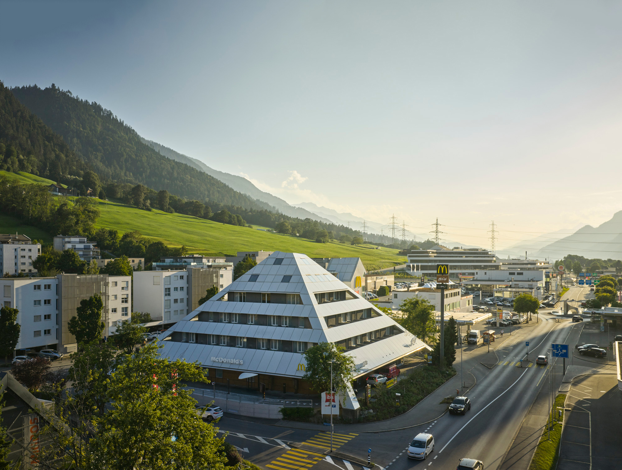 Gebäude des Ibis Hotels in Chur mit pyramidenförmiger Architektur, Straße mit Autos im Vordergrund und grünen Hügeln sowie Bergen im Hintergrund bei Sonnenuntergang.