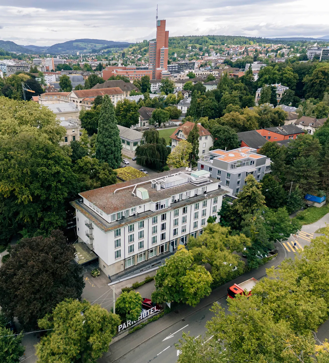 Luftaufnahme des Park Hotels in Winterthur, umgeben von grünen Bäumen, mit Stadt und Hügeln im Hintergrund unter bewölktem Himmel.