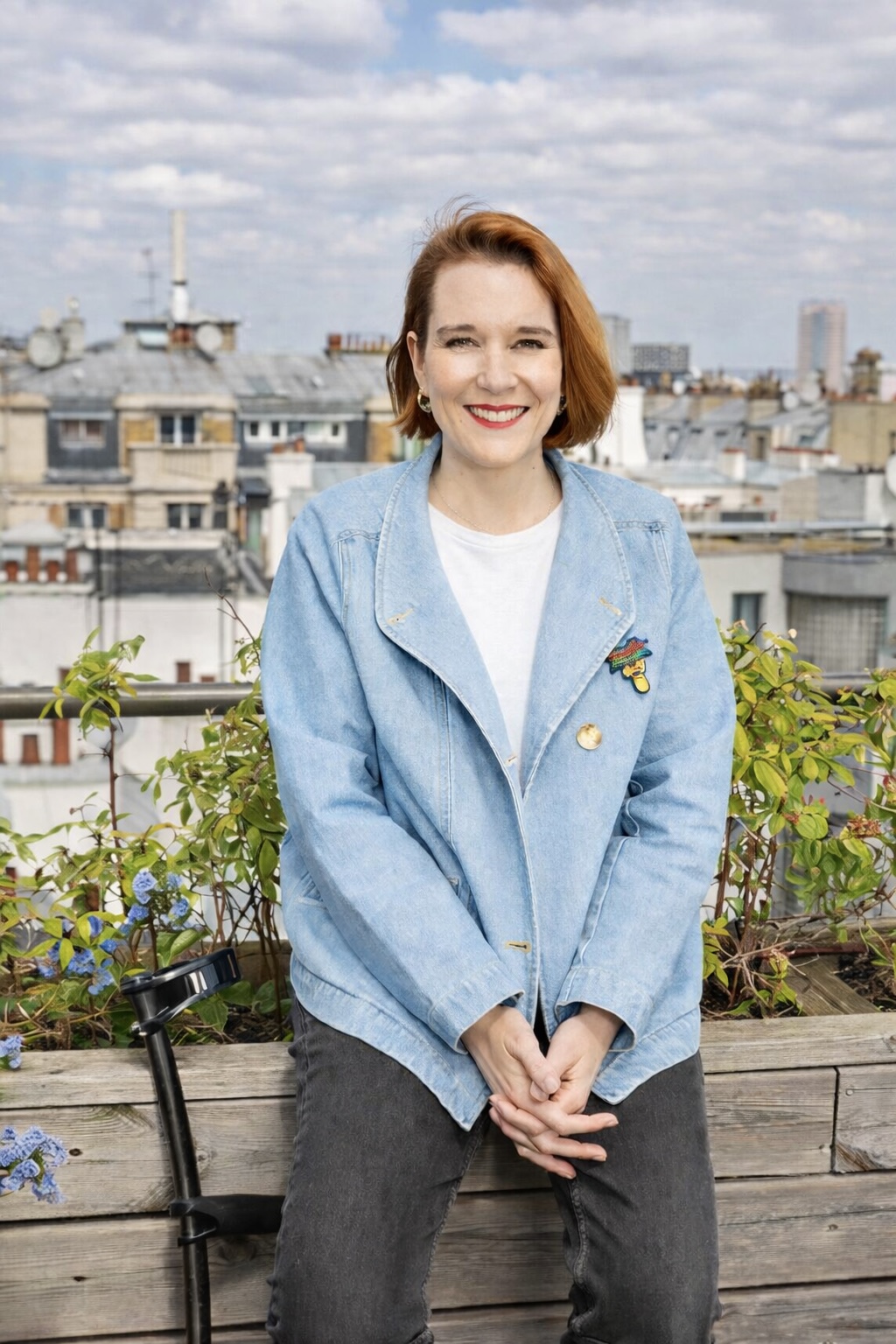 Femme souriante aux cheveux roux portant une veste en jean bleue et un jean noir, assise sur un banc en bois avec un paysage urbain en arrière-plan.