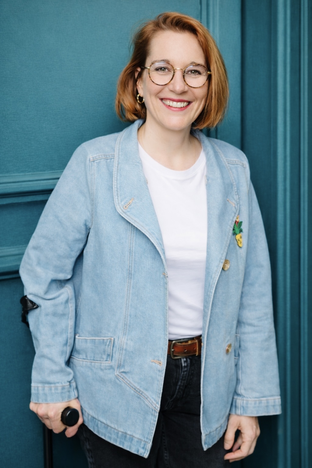 Femme souriante à lunettes avec veste en jean bleu clair, t-shirt blanc et jeans noirs, posant devant un mur bleu.
