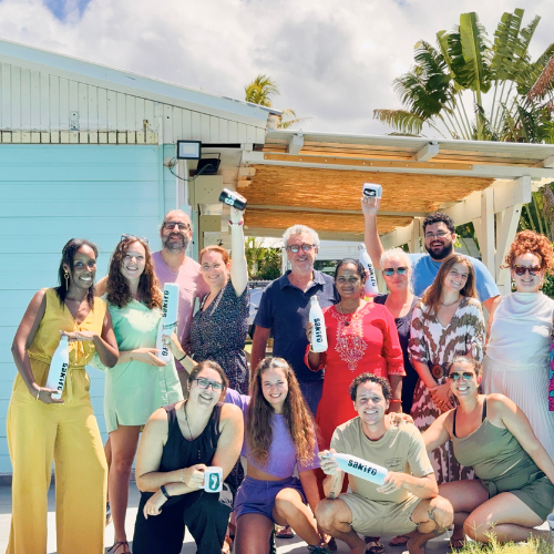 Groupe de personnes souriantes en extérieur, certaines tenant des bouteilles blanches avec des logos, devant une maison avec végétation tropicale.