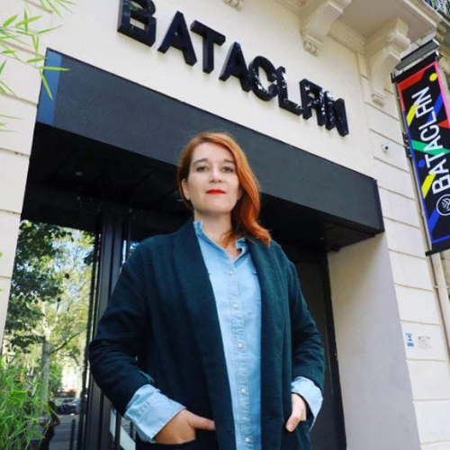 Femme aux cheveux roux portant un manteau bleu et une chemise, posant devant l'entrée du Bataclan à Paris.