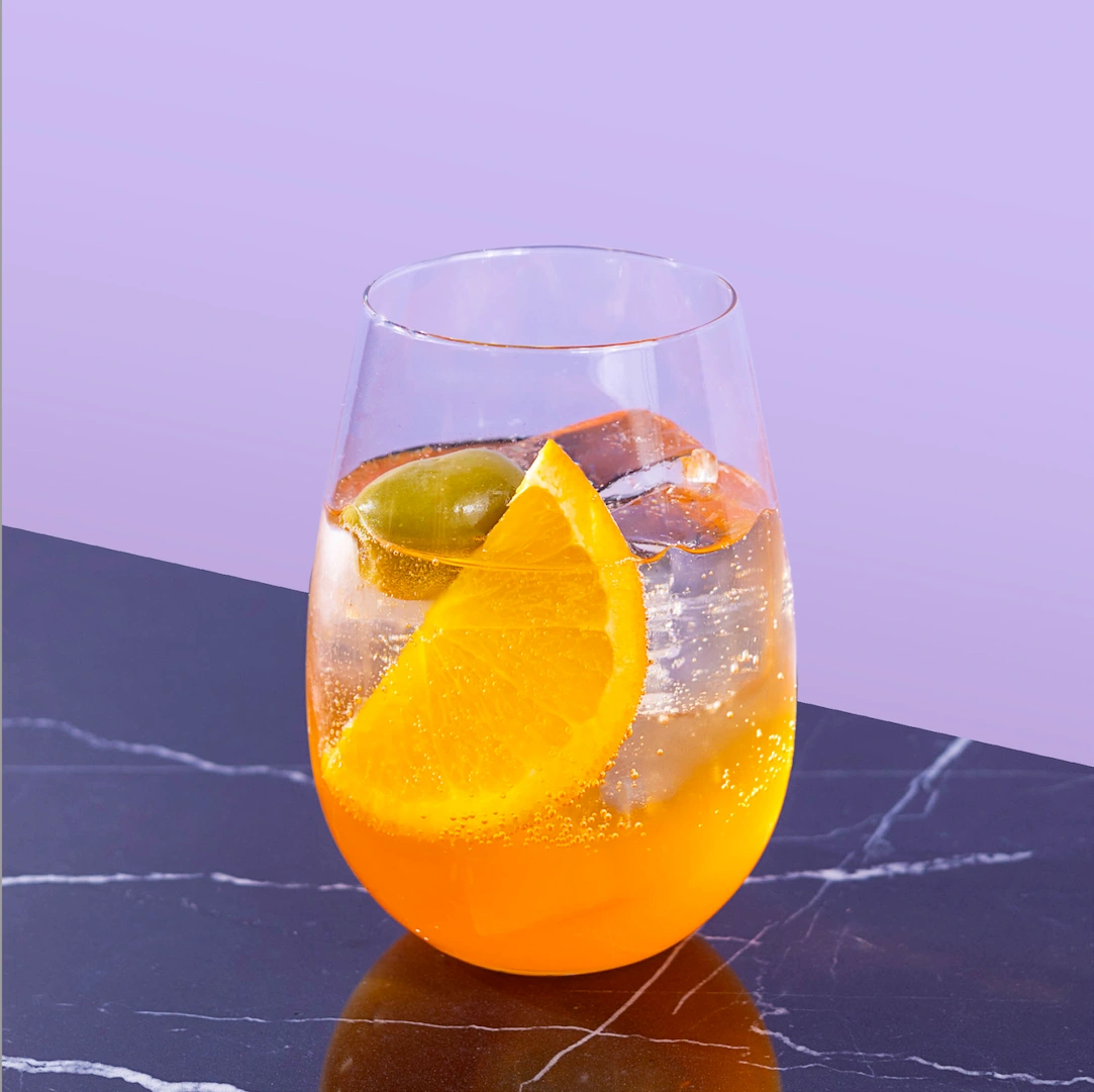 Cocktail à base d'apéritif italien, de mousseux et d'eau pétillante sur fond mauve.