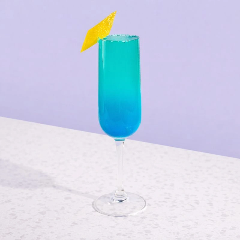 Cocktail made with blue curaçao, lychee syrup, lemon and sparkling wine on a lilac background.