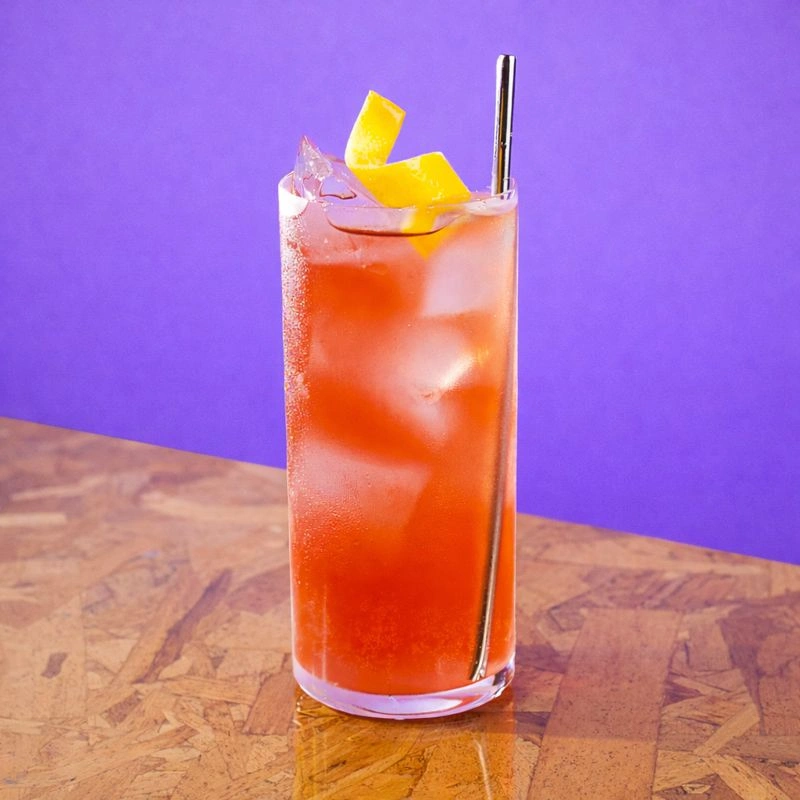 Cocktail à base de gin, de sirop de tonic à l'hibiscus et d'eau pétillante sur un fond mauve.