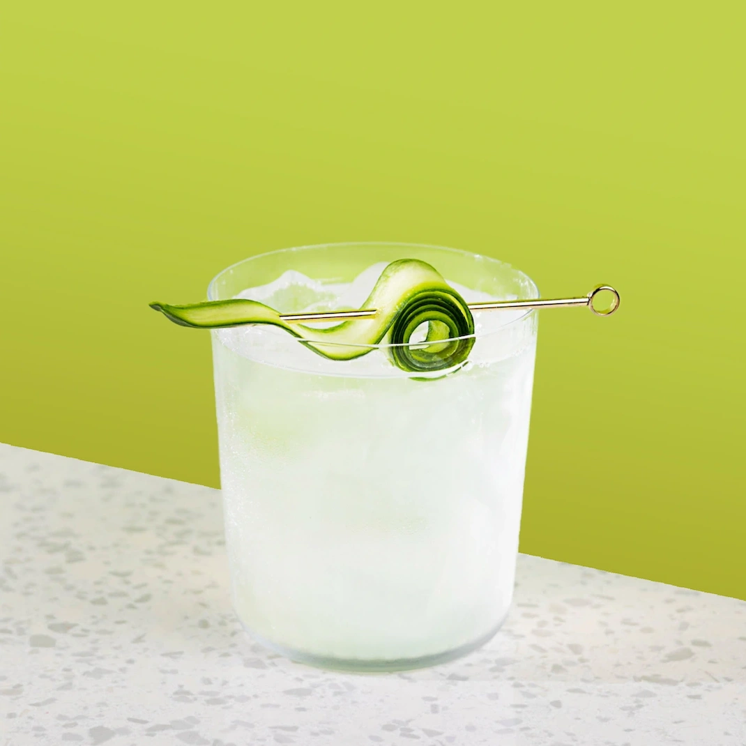Cocktail à base de gin, de sirop de lime, de concombre et de soda tonique sur un fond vert.