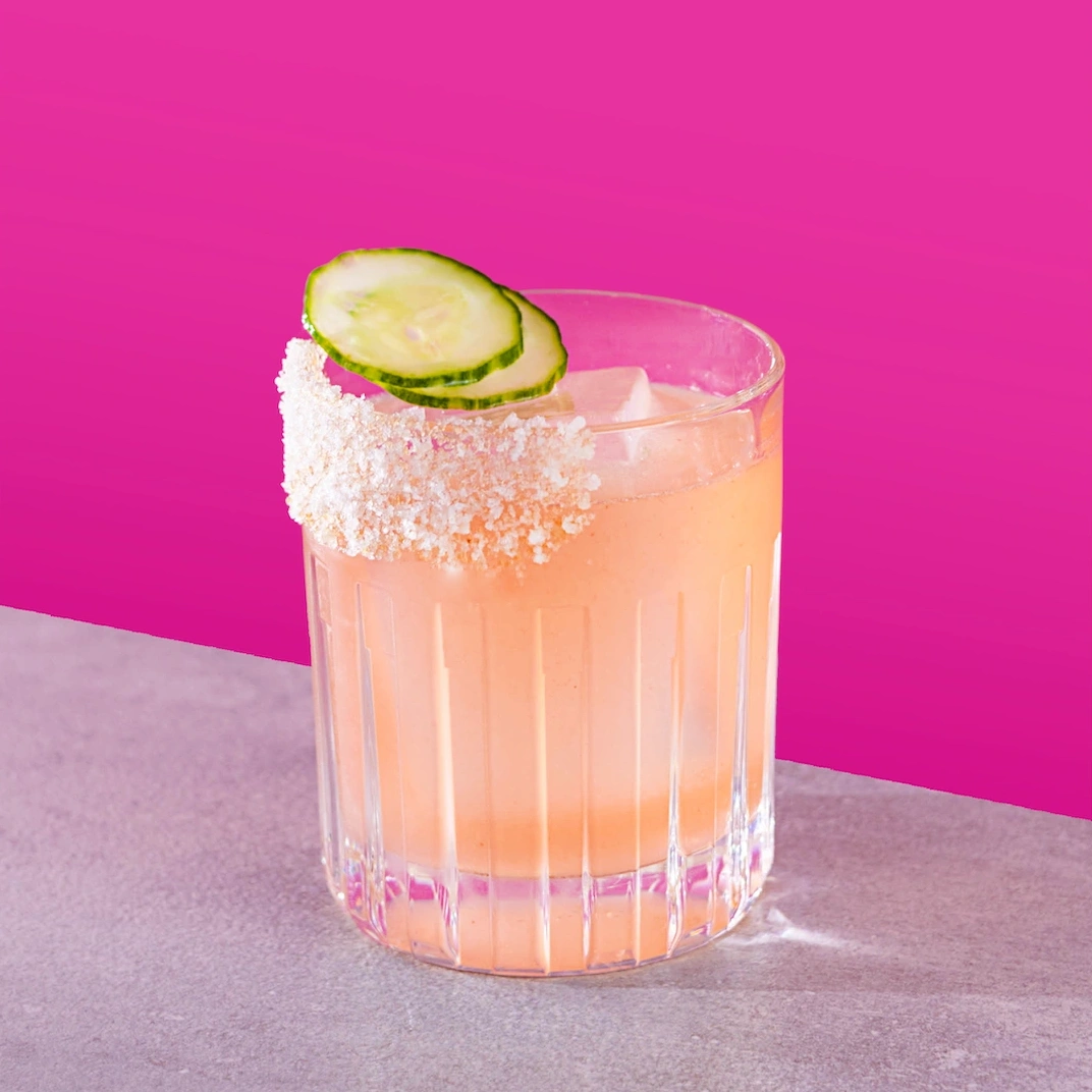 Cocktail à base de tequila, de sirop de licorne rose, de jus de lime frais et de concombre sur fond fushia.