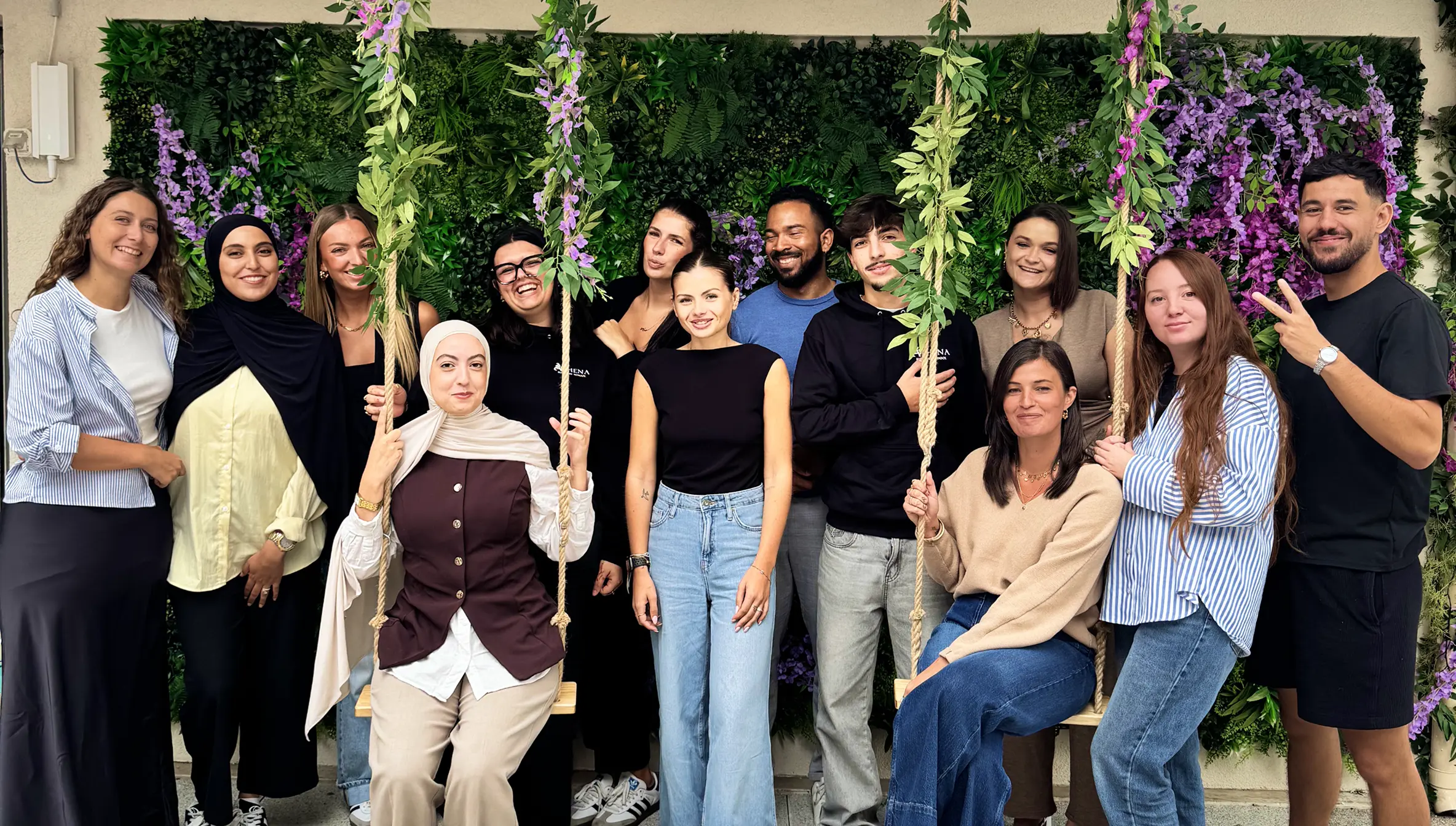 Groupe de 13 personnes souriantes posant devant un mur végétal avec des balançoires décorées de fleurs violettes.
