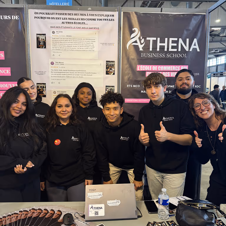 Groupe d'étudiants souriants portant des sweat-shirts noirs Athena Business School devant un stand d'information avec une affiche et un ordinateur portable.