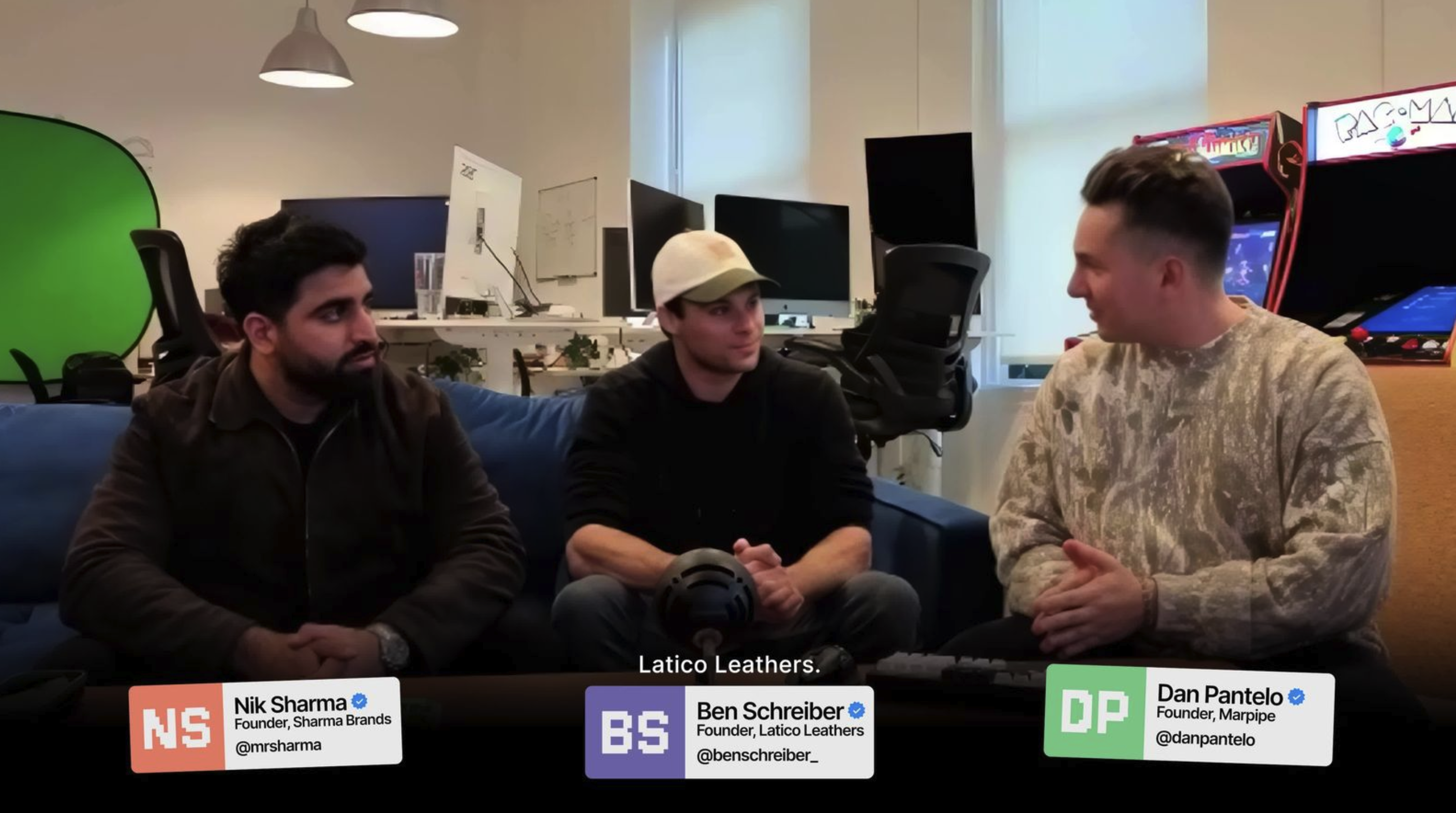 Three men sitting on a blue couch in an office with computers and arcade machines behind them, engaged in conversation.