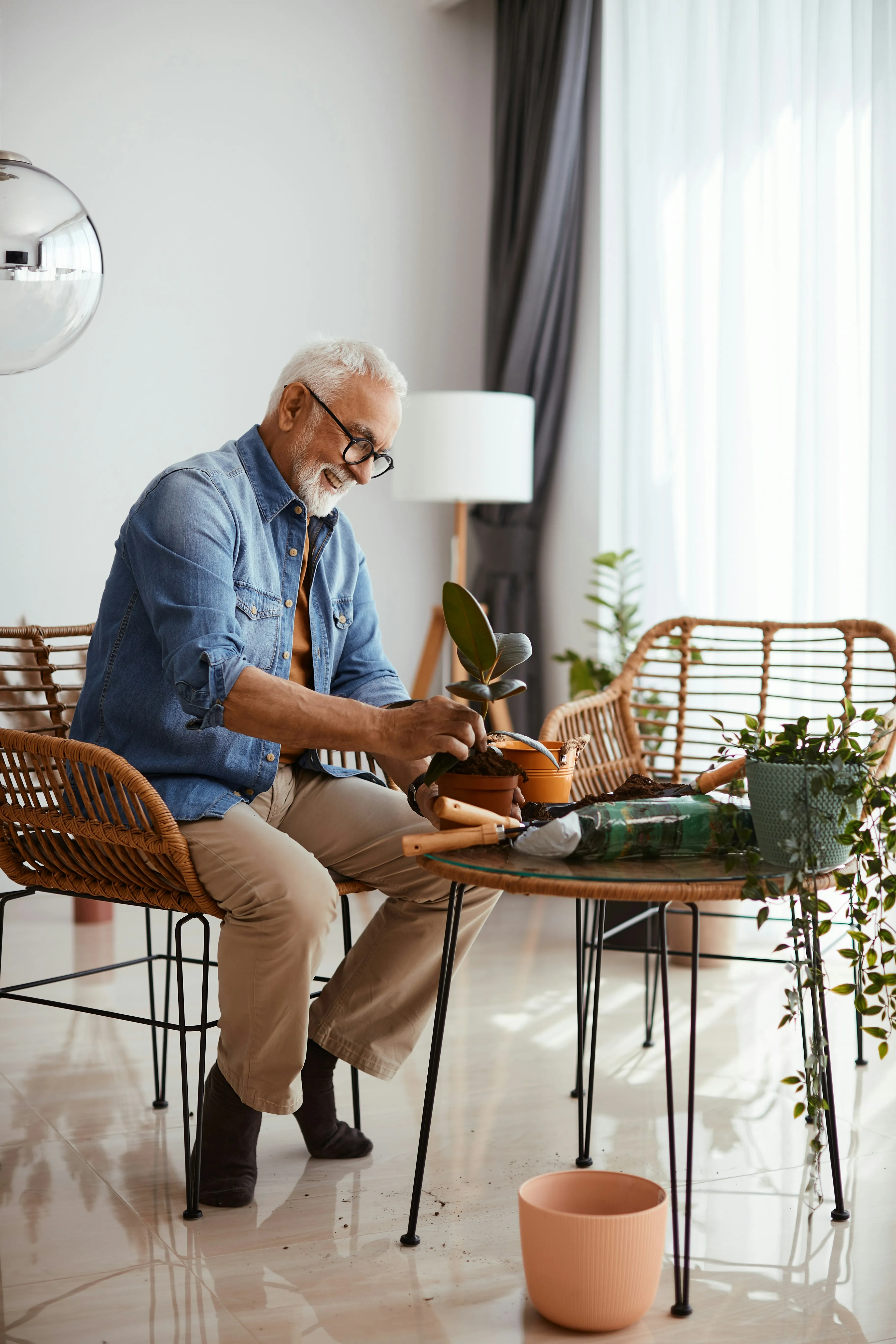 Elderly man in glasses and denim shirt potting a plant indoors at a table with gardening tools and soil.