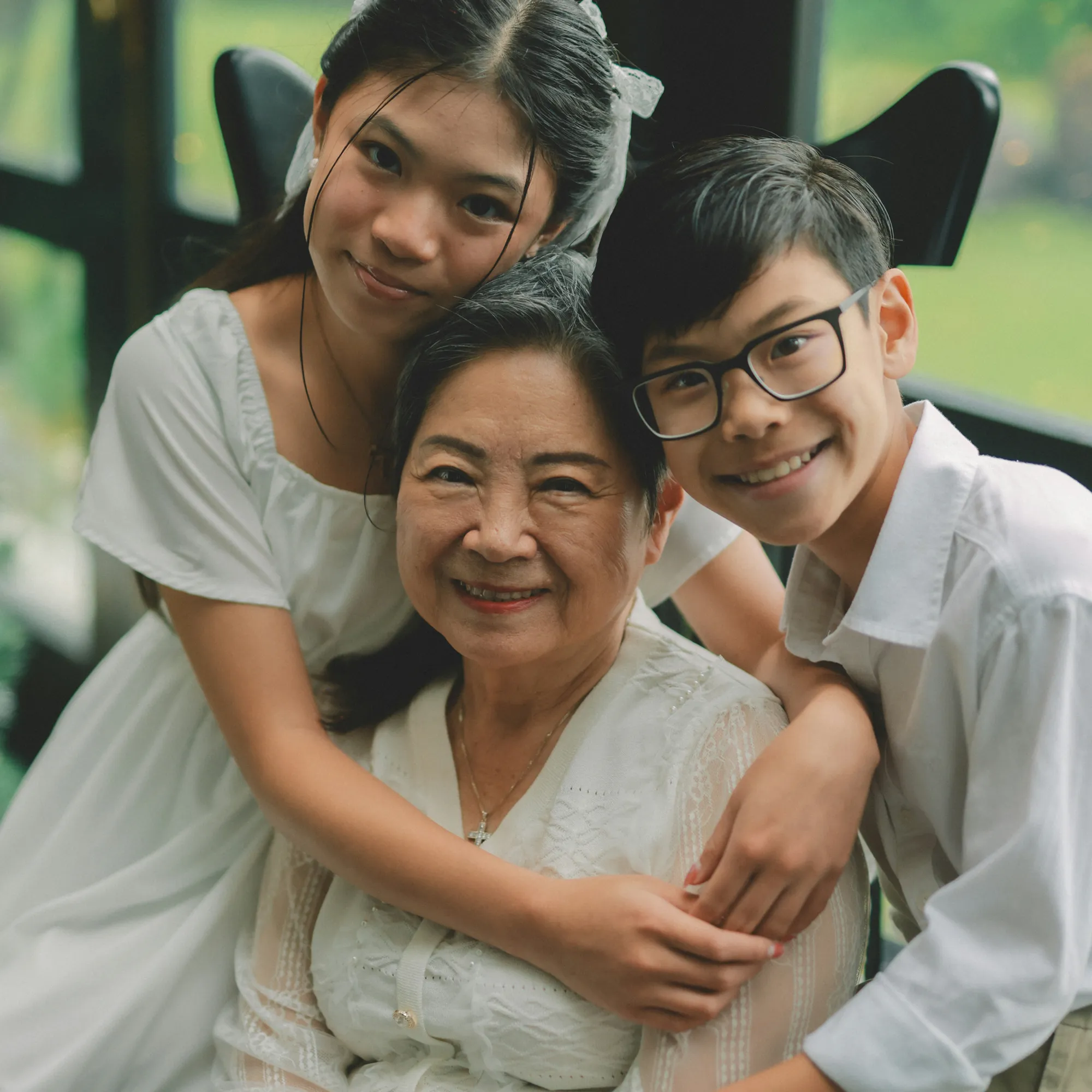 Smiling elderly woman embraced by a young girl and a boy indoors near a window.