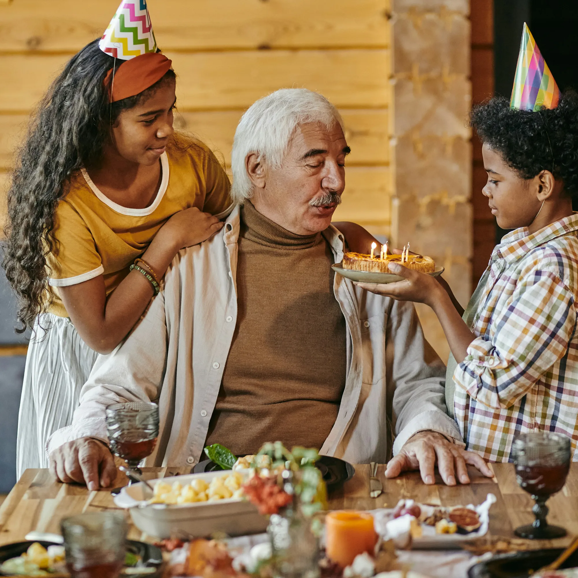 Grandfather wearing a beige shirt blowing out candles on a birthday cake held by his grandson, while granddaughter wearing a party hat stands beside him.