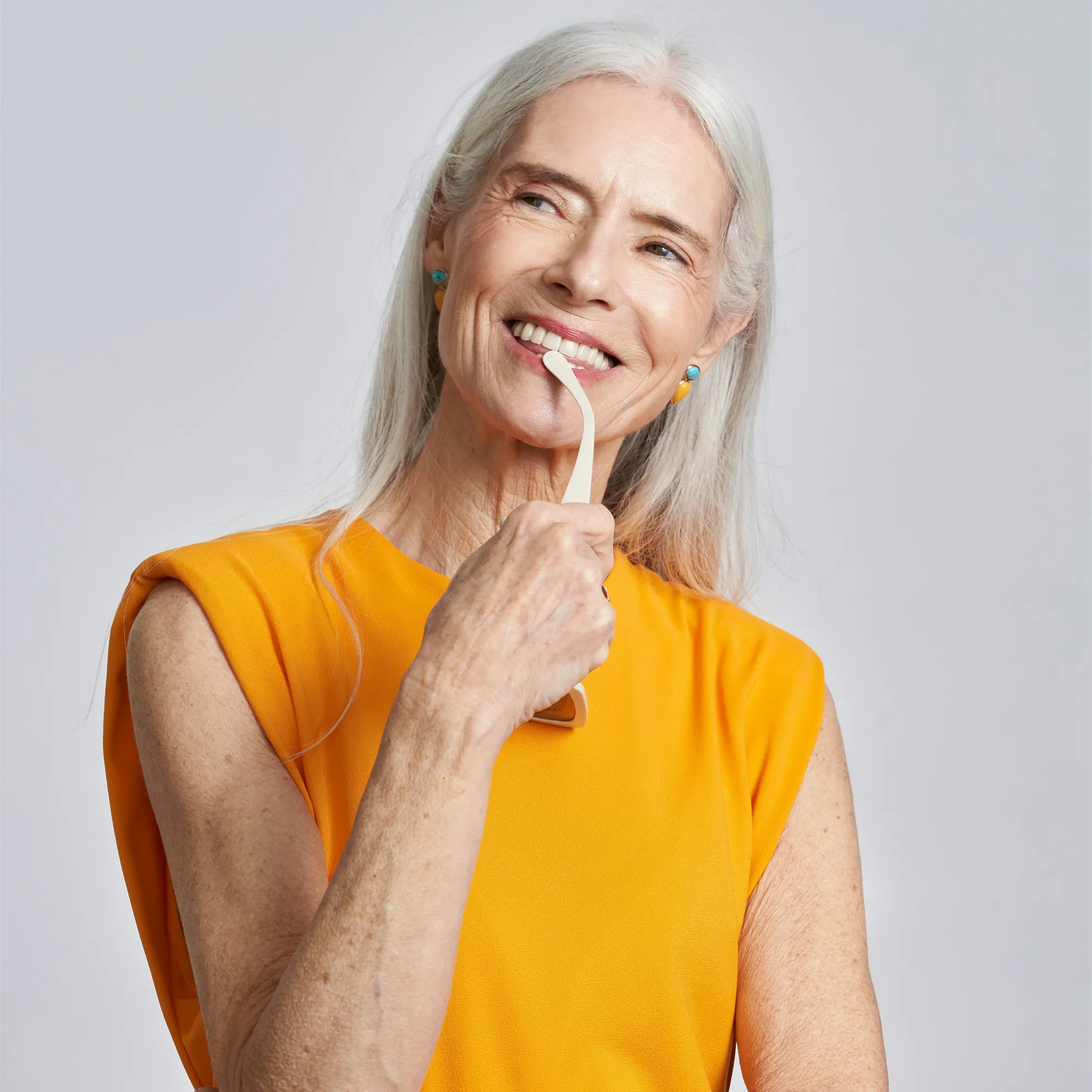 Smiling elderly woman with long white hair wearing an orange sleeveless top, holding a beige facial roller near her chin.