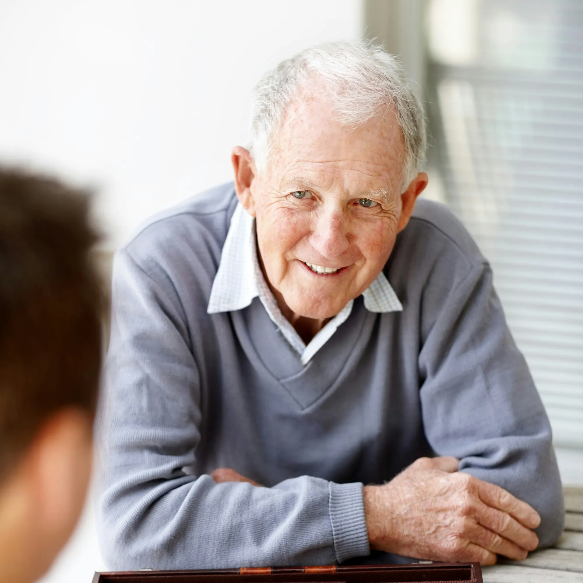 Smiling elderly man with gray hair wearing a gray sweater sitting at a table with a backgammon board.