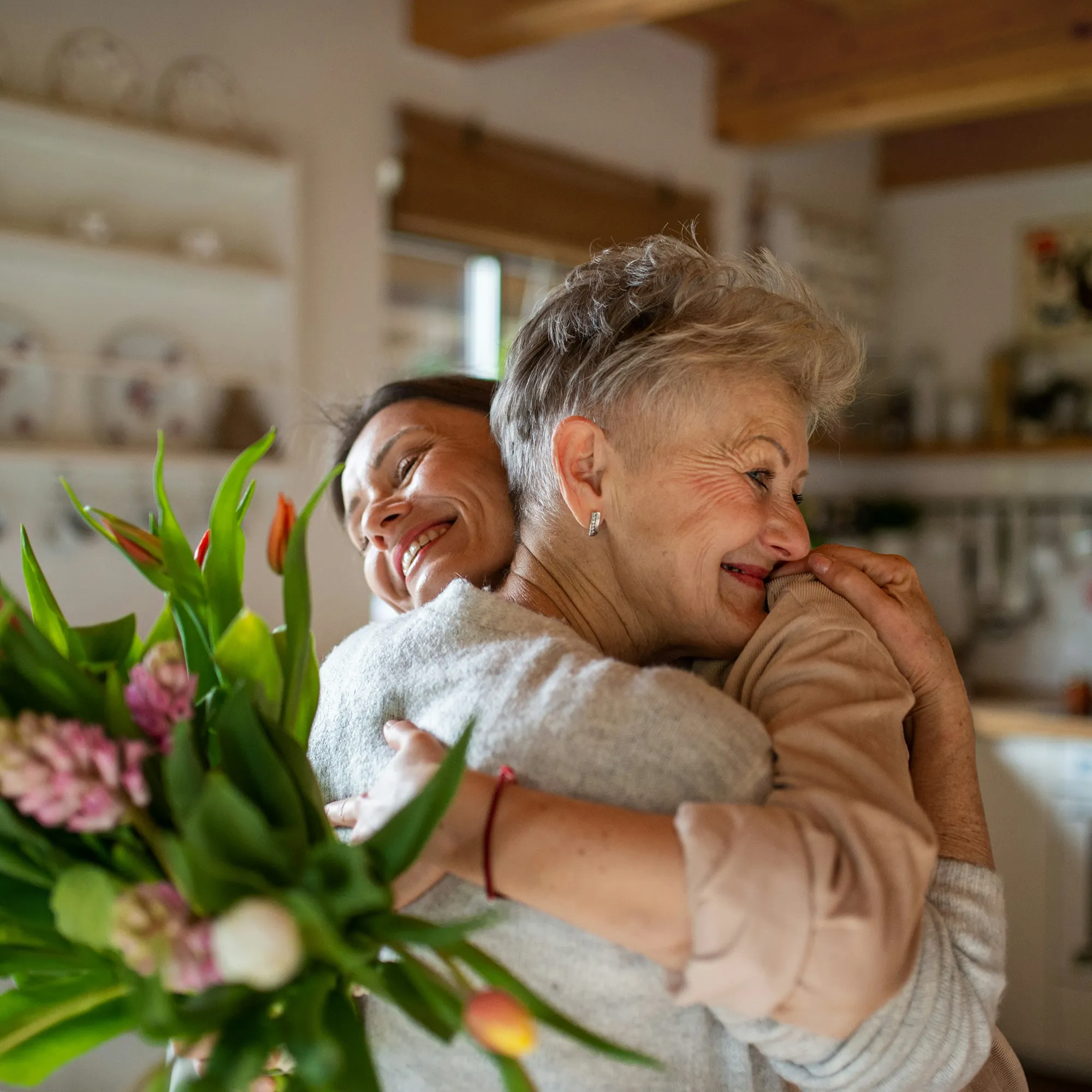 Two women sharing a warm hug indoors, one holding a bouquet of flowers and both smiling happily.