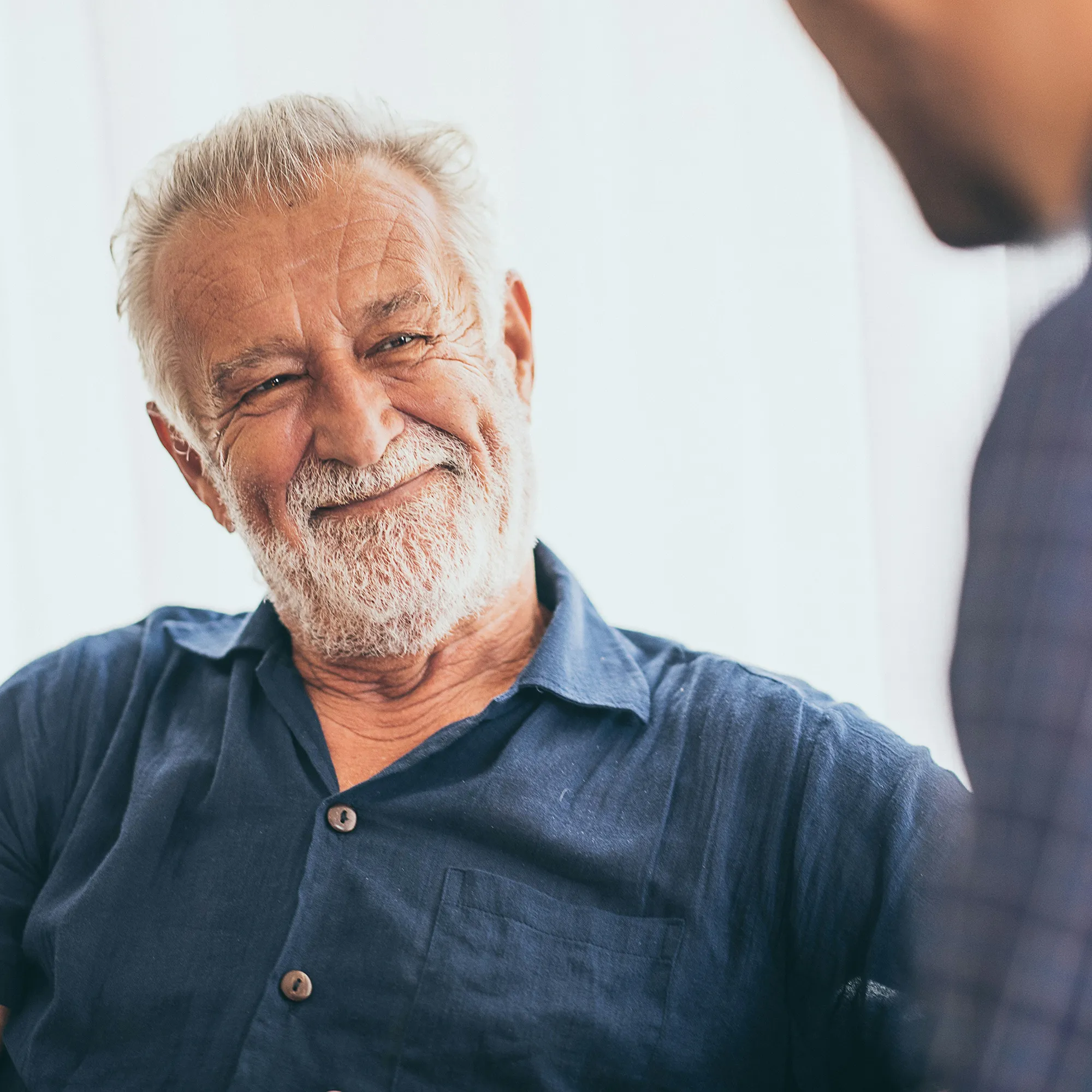 Smiling elderly man with white hair and beard wearing a blue shirt, engaged in conversation.